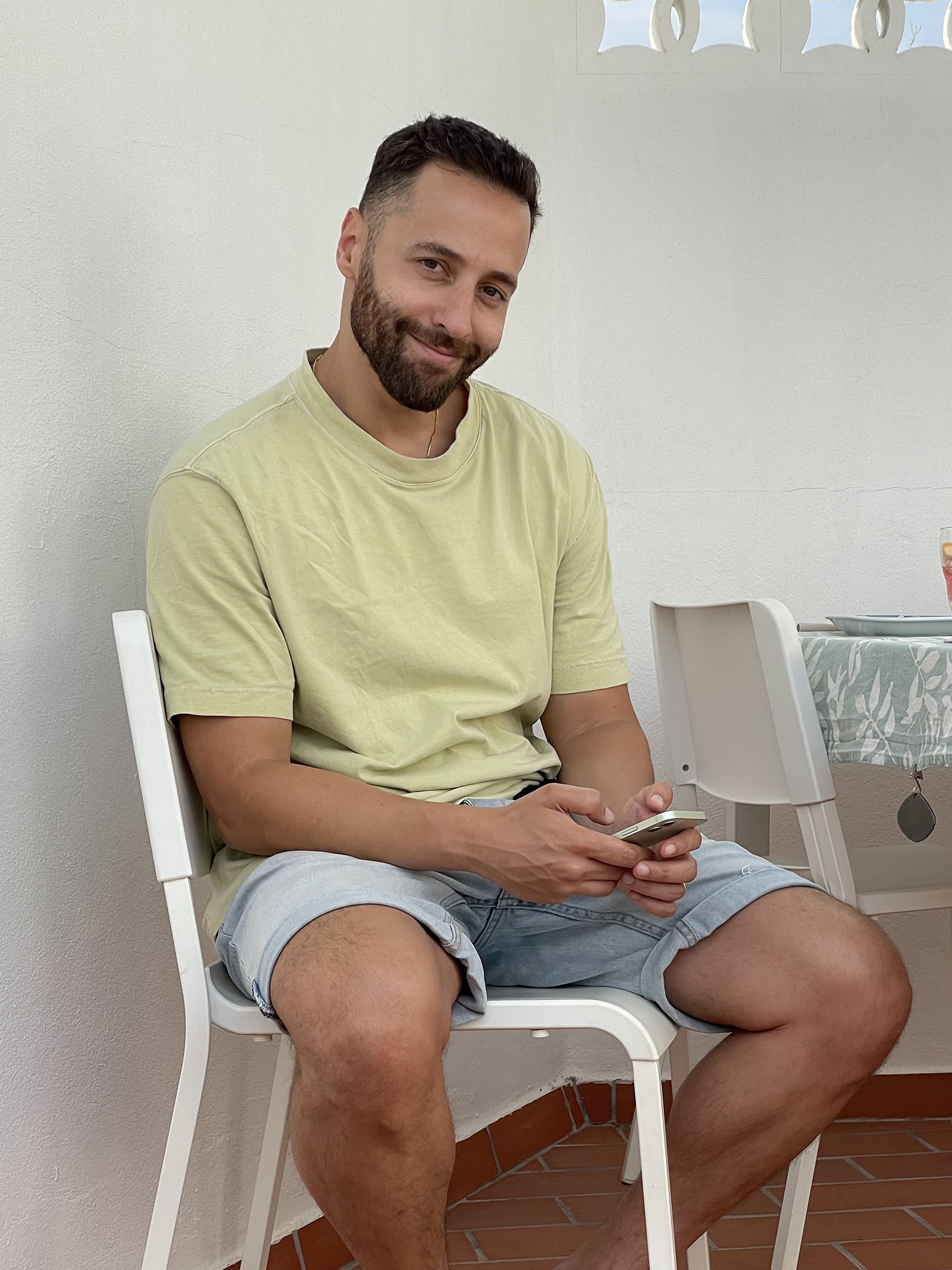 A man with dark hair and a beard, wearing a yellow t-shirt and light denim shorts, sitting on a white chair, smiling and holding a smartphone. He is indoors, against a white wall, with a table and chair nearby.