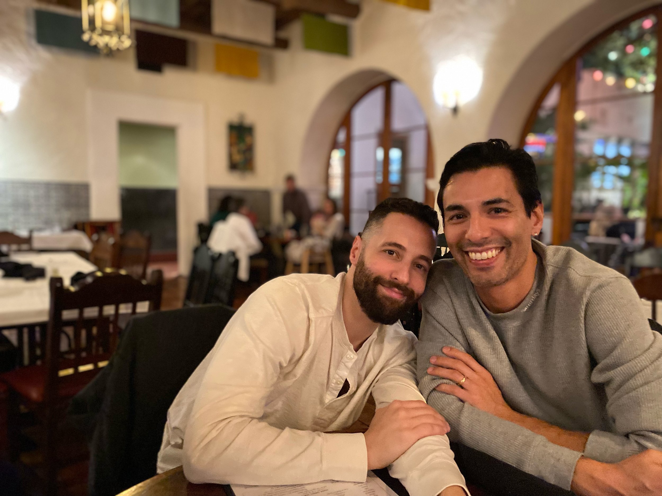 Two men smiling and sitting at a restaurant table in a cozy, warmly lit dining area with other patrons in the background.