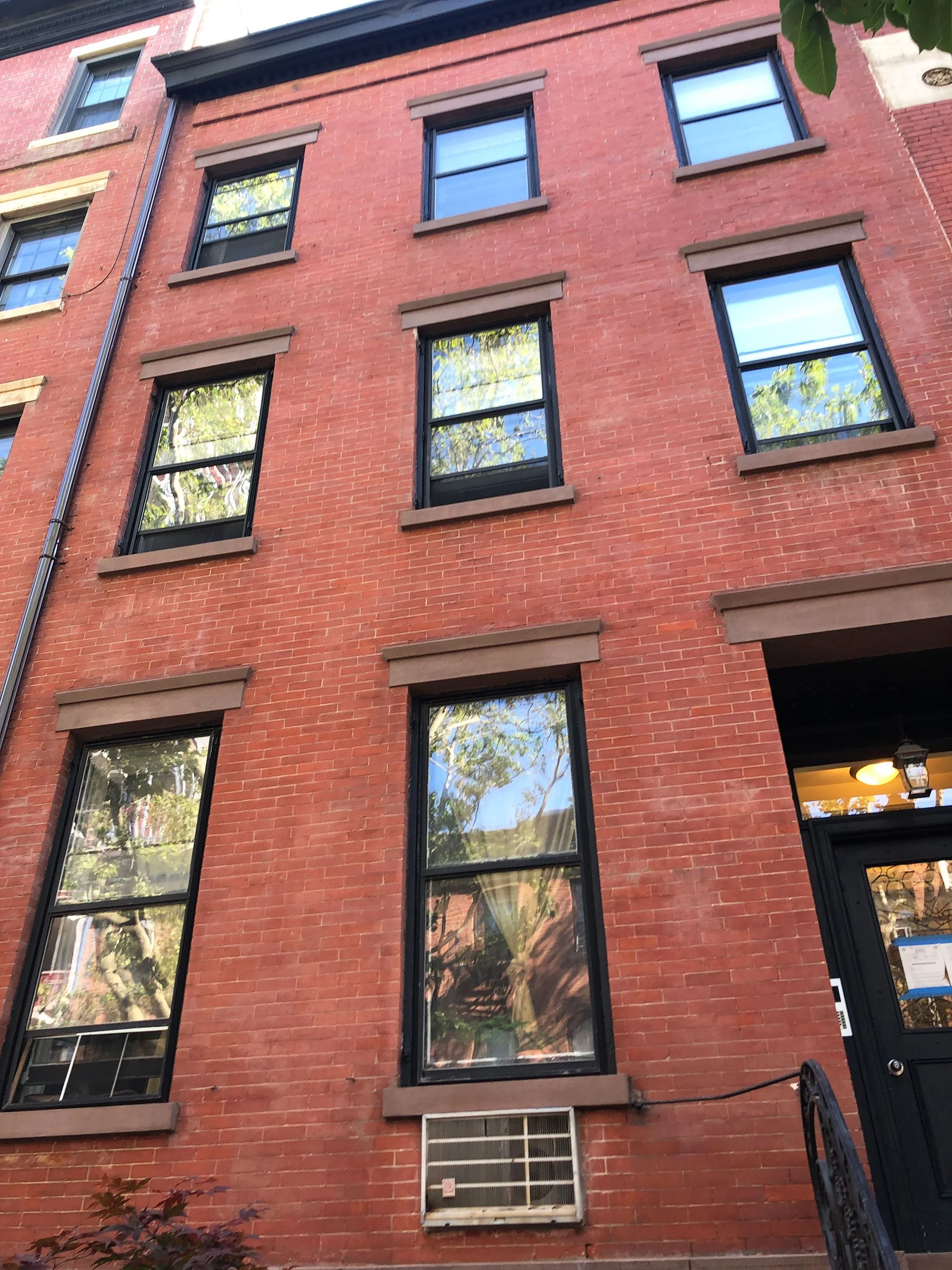 Red brick apartment building with multiple black-framed windows reflecting trees, with a small air conditioning unit below one window, and a staircase with a black railing leading to the entrance