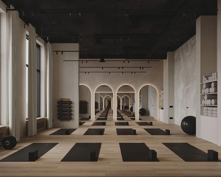 Empty yoga studio with black mats and blocks, large windows with white curtains, mirrored arches, and exercise balls.