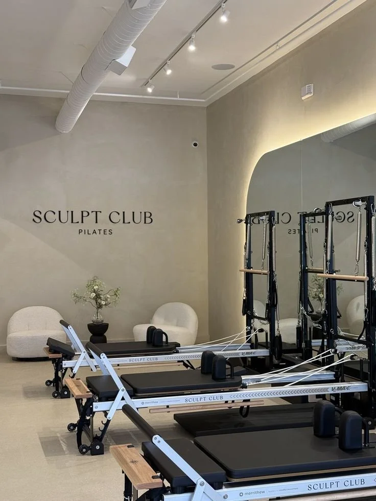 Interior of a Pilates studio with reformer machines, a beige wall with 'SCULPT CLUB PILATES' signage, and comfortable chairs with a small plant.