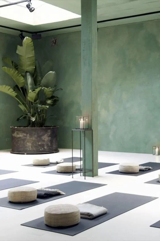 Yoga studio with mats and bolsters on the floor, a large potted plant, and candles on a stand, with minimalist decor and a green painted wall.