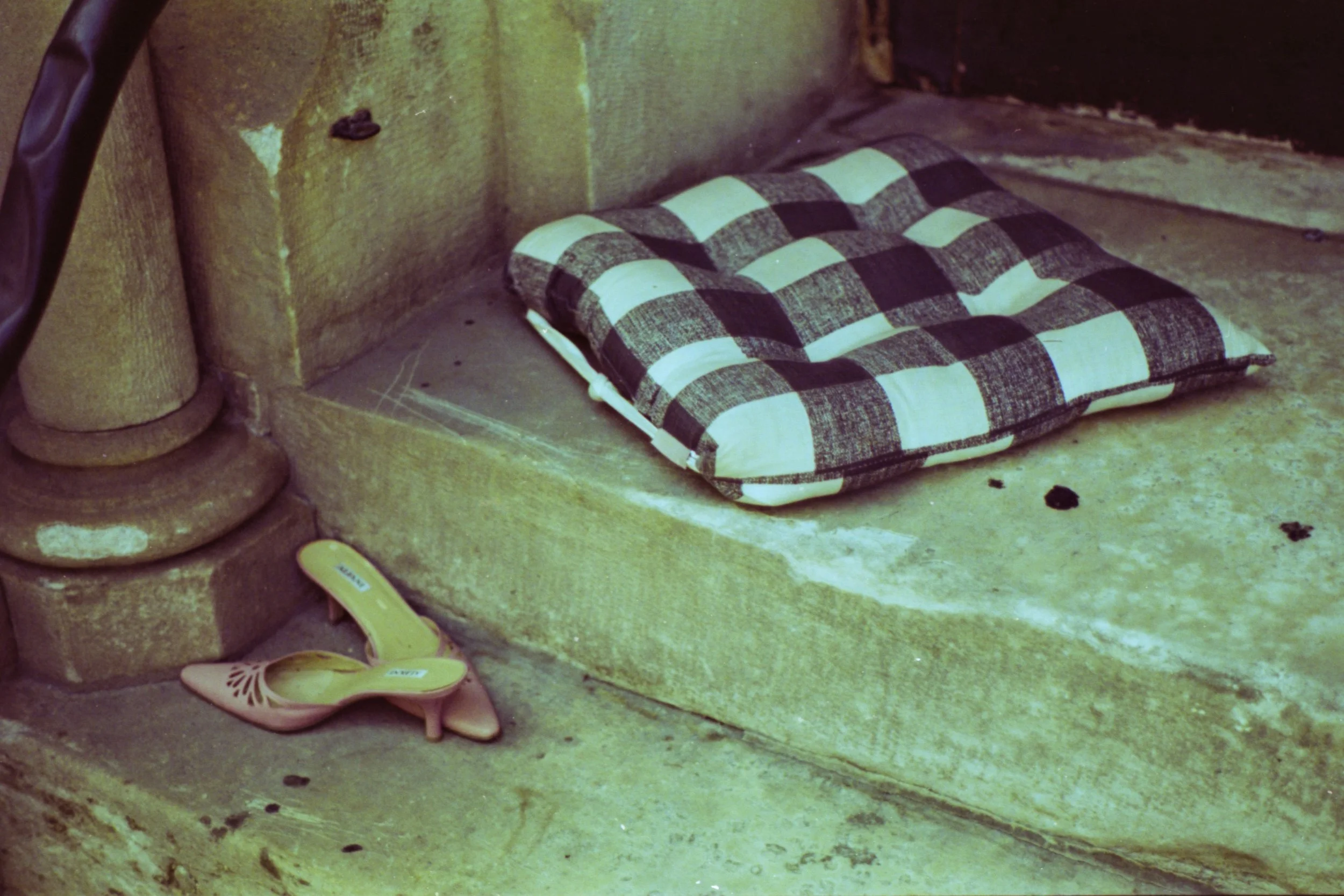 A pair of pink high-heeled shoes and a plaid cushion are placed on a stone step near a corner of an old, weathered building.