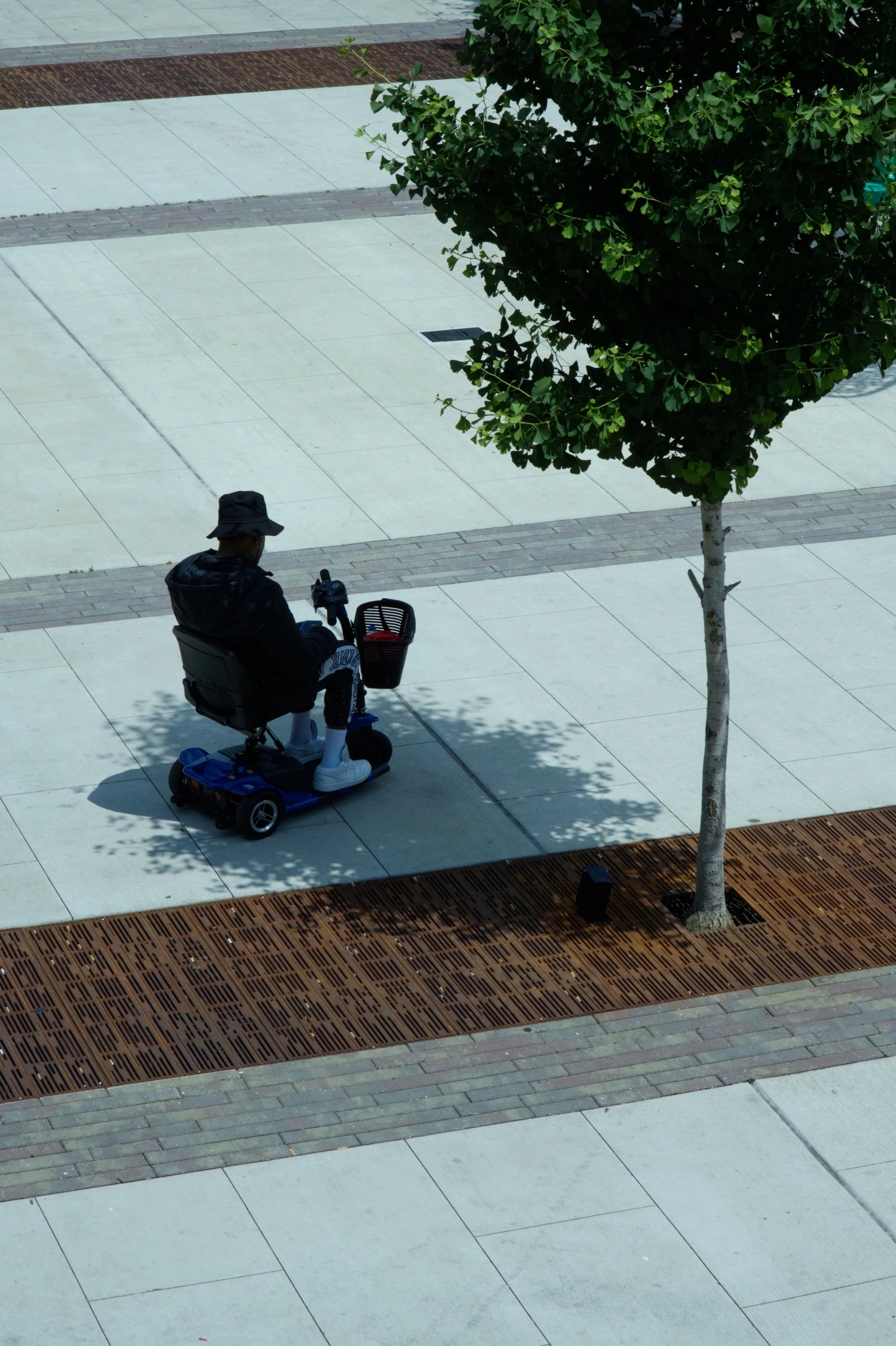 A man riding a mobility scooter along a paved outdoor area with a tree and shadows.