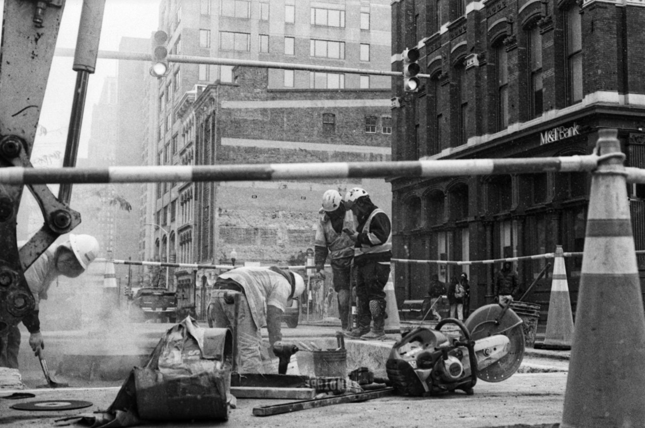 Construction workers on a city street working with tools, surrounded by safety barriers, with tall buildings and a bank in the background.
