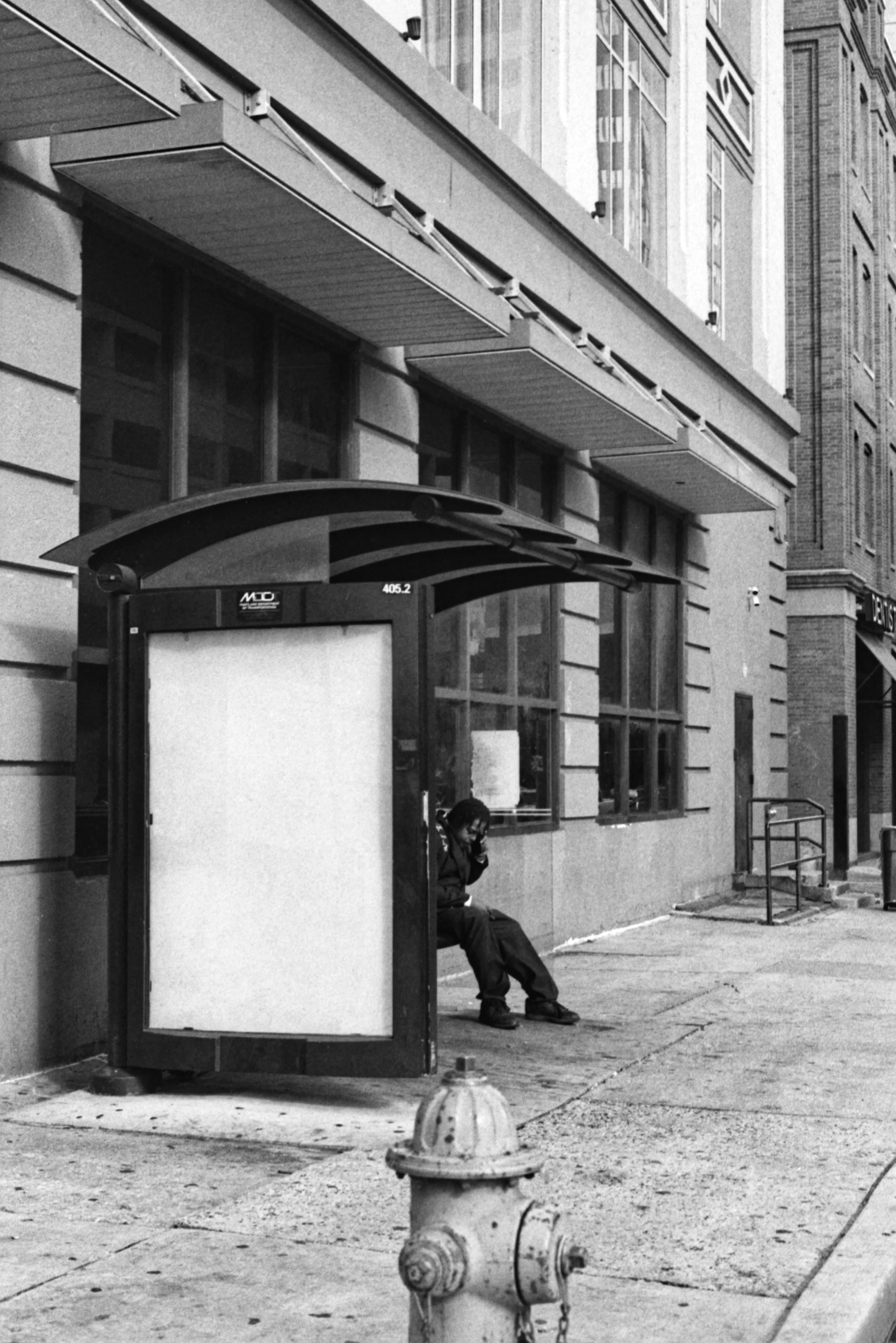 A person sitting at a bus stop on a city sidewalk, talking on the phone, with a fire hydrant nearby and tall buildings in the background.