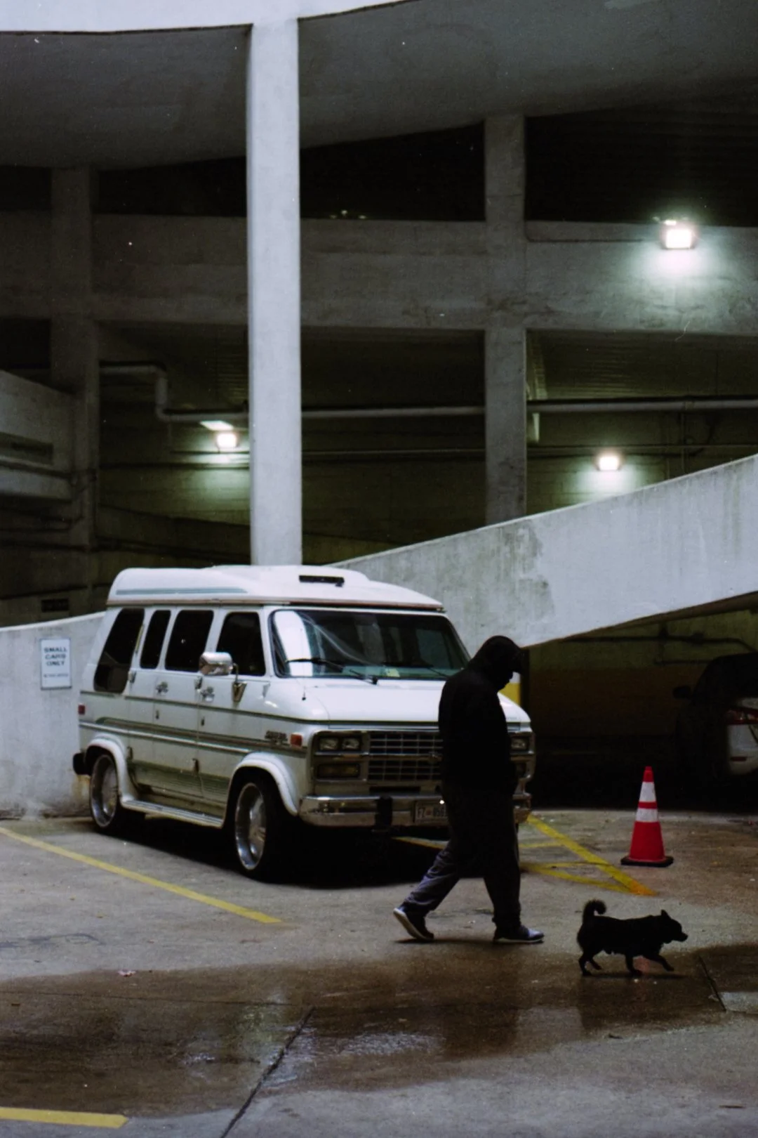 A person wearing a black hoodie walking a small black dog in a parking garage at night. There is an old white van and some parked cars, with concrete parking structure and dim lighting.