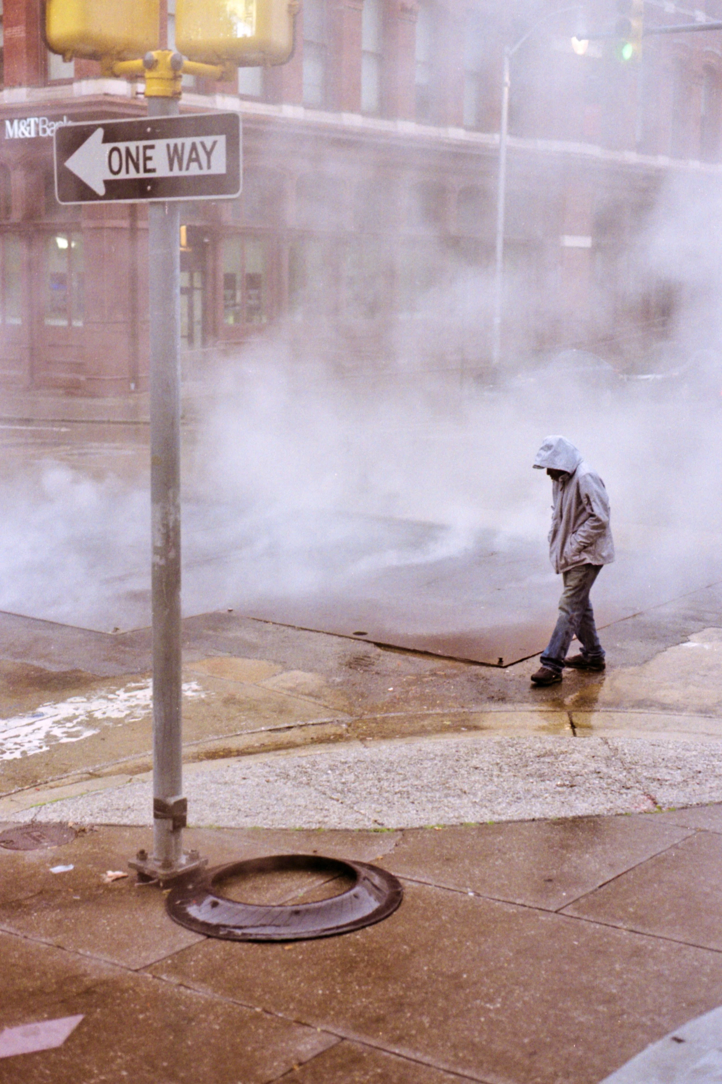 A person in a gray hoodie and jacket walking on a city sidewalk near steam rising from a manhole cover, with a one-way street sign pointing left in the foreground.