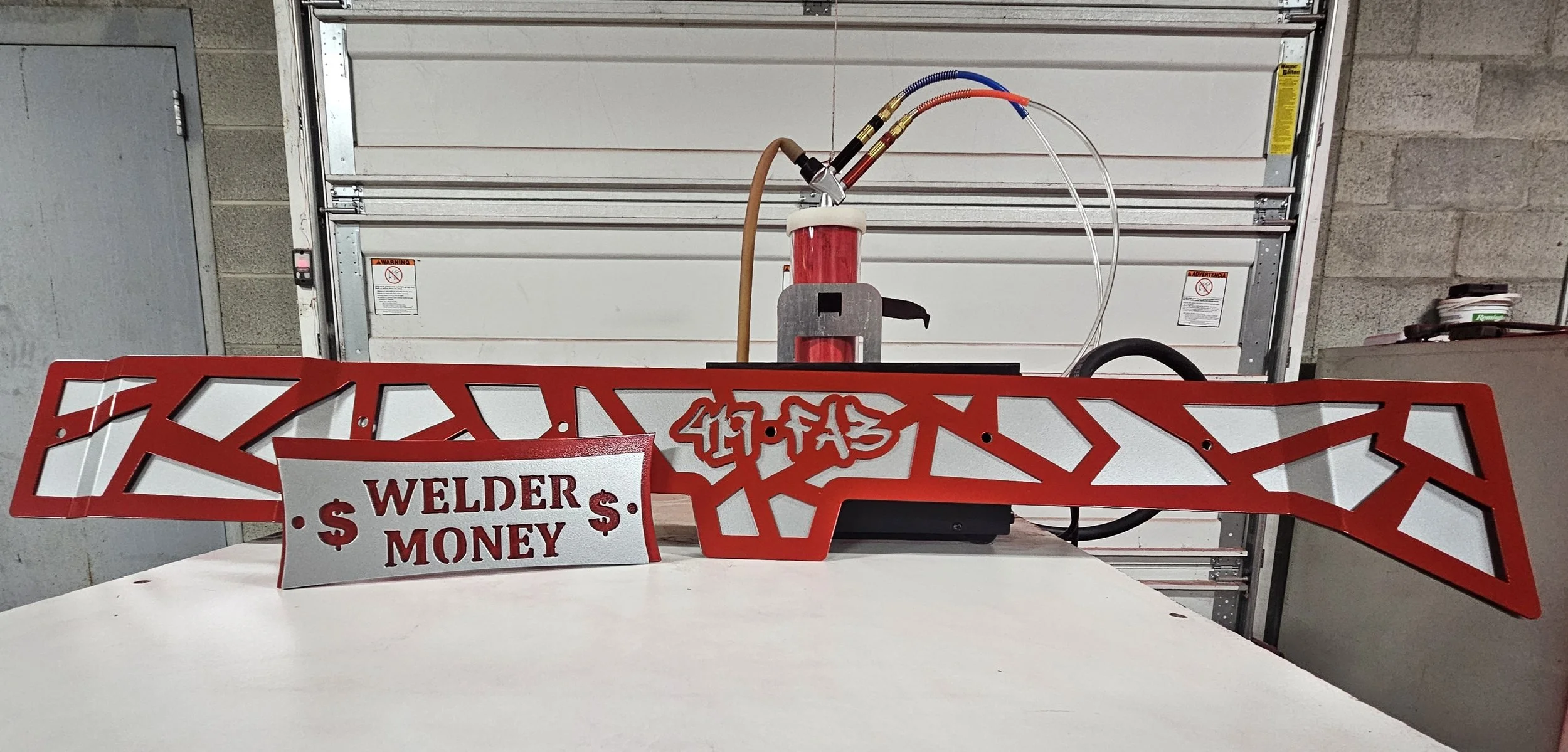 Red metal structure with graffiti and signs, labeled 'Welder Money' on a white table in an industrial setting.