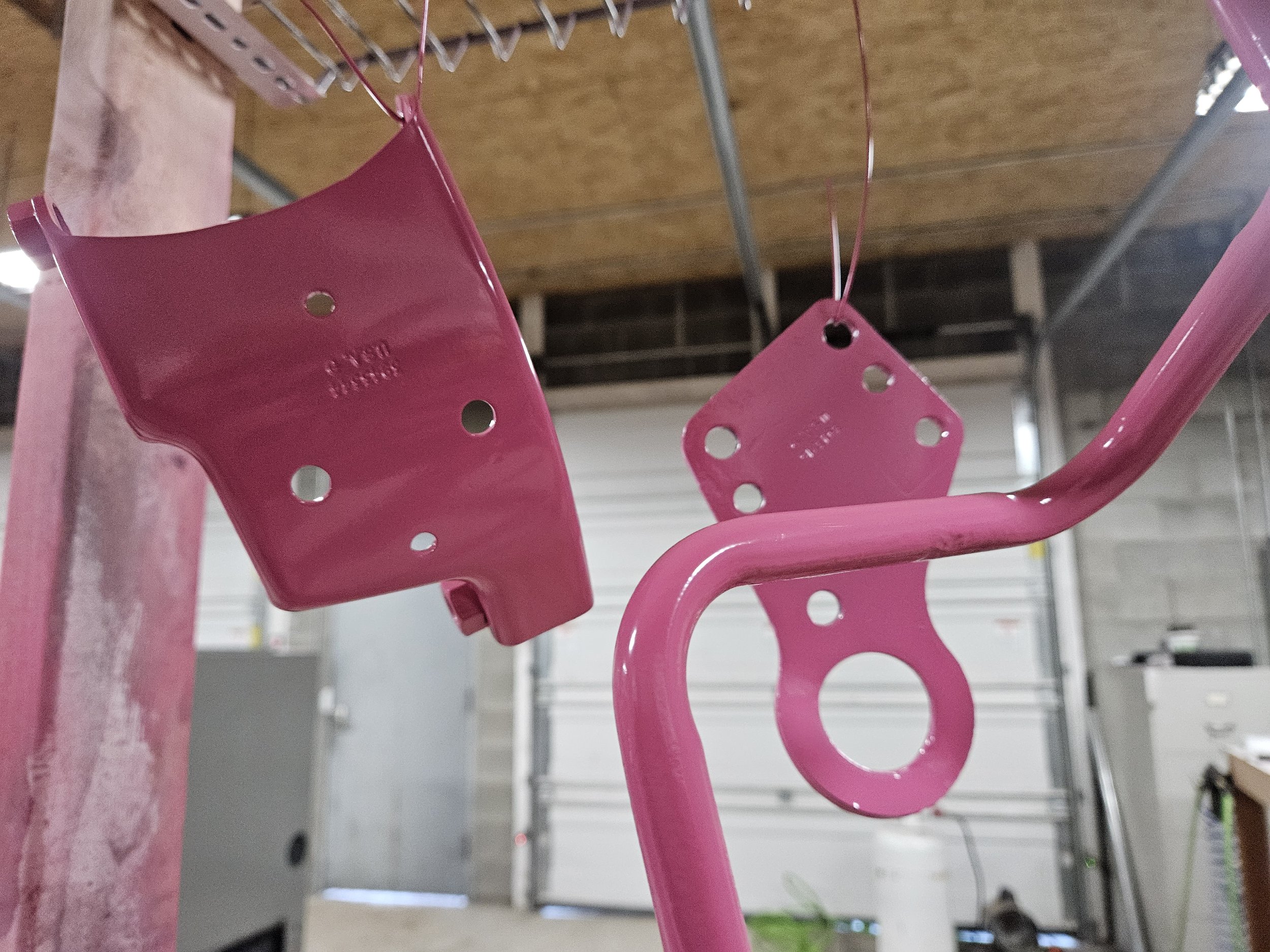 Pink metal components hanging from wires, likely parts of a bicycle or similar assembly, in a workshop setting.