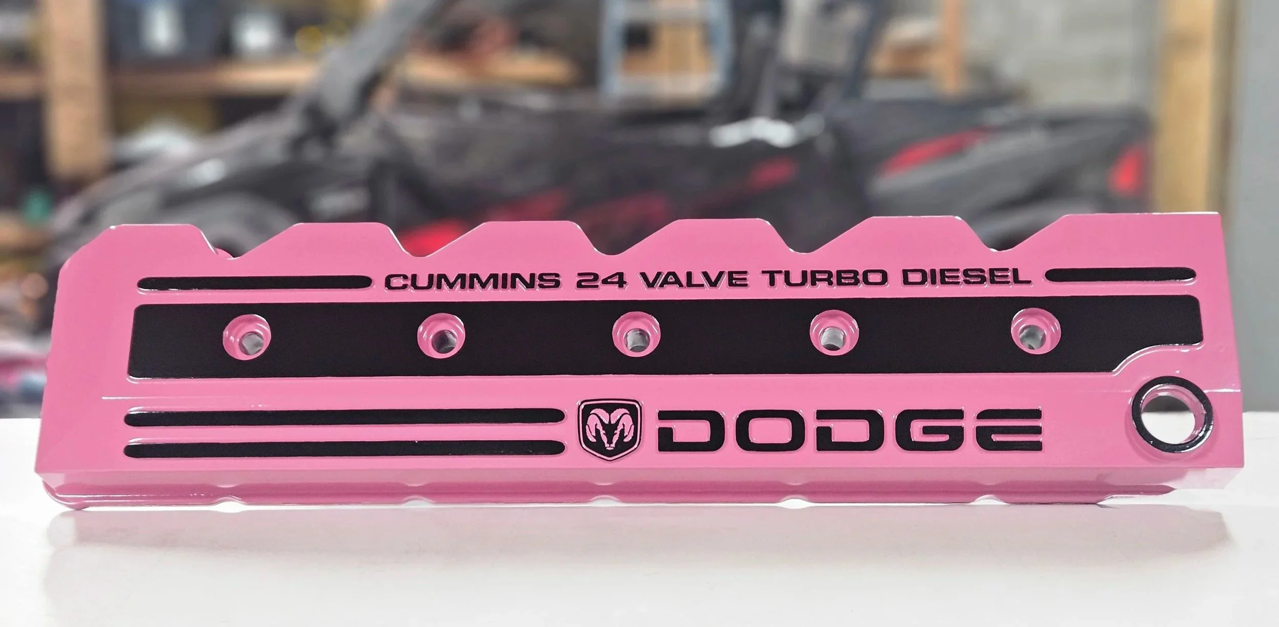 Pink valve cover with black accents and the logo of Dodge, labeled as a Cummins 24 valve turbo diesel engine part, placed on a white surface with a blurred background.
