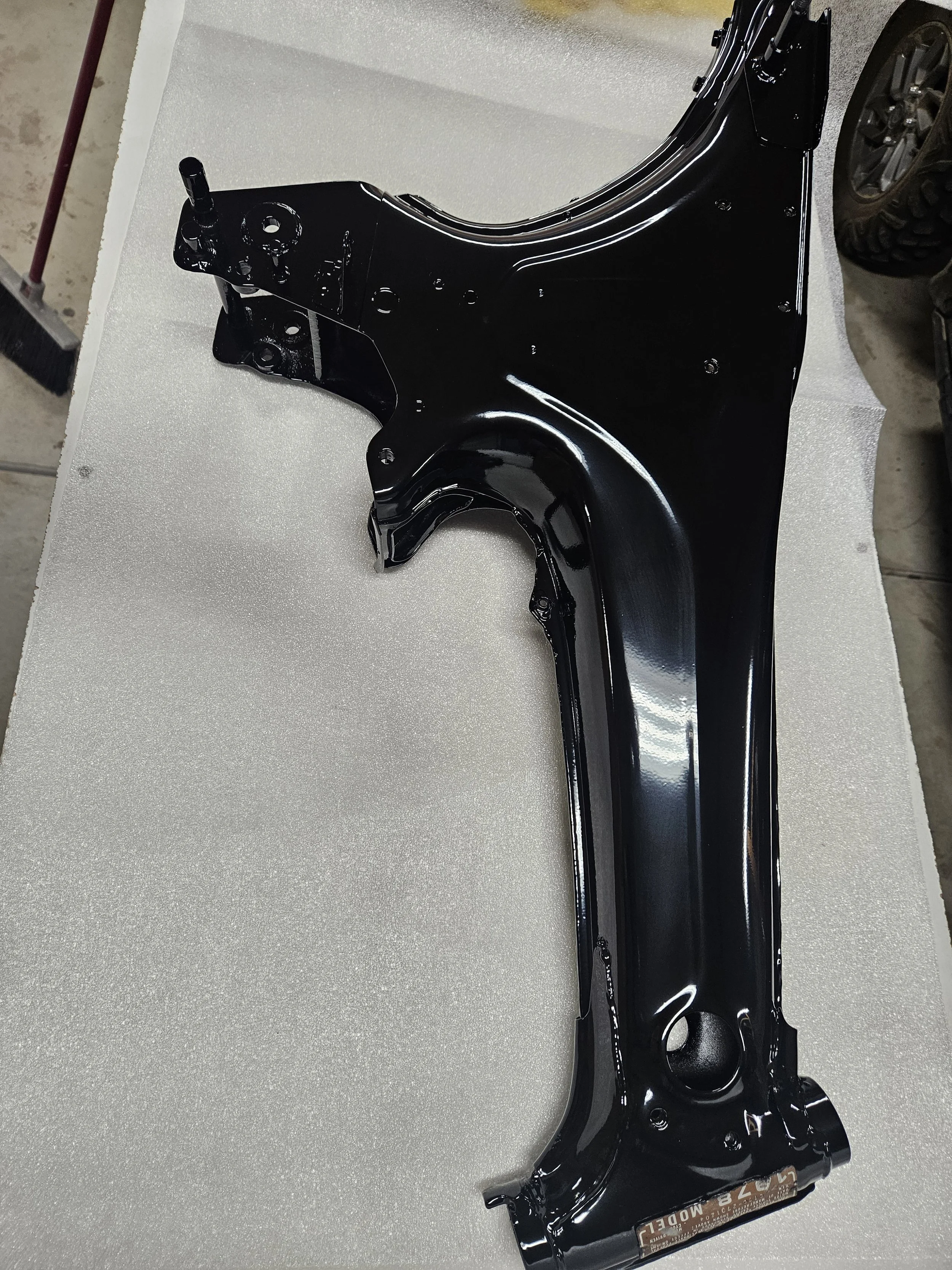 A black painted metal car part, possibly a dashboard frame or chassis component, lying flat on a foam surface in what appears to be a workshop.