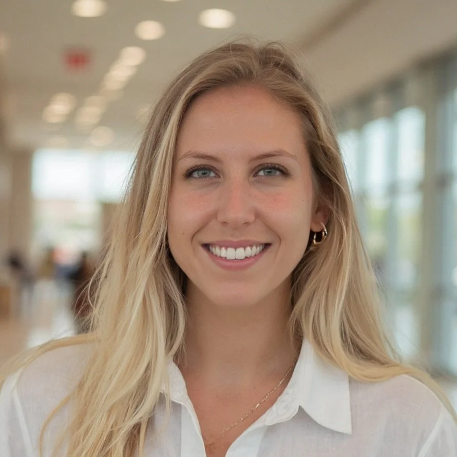 A young woman with long blonde hair and light skin, smiling, wearing a white shirt and small hoop earrings, standing in a bright indoor space with large windows.