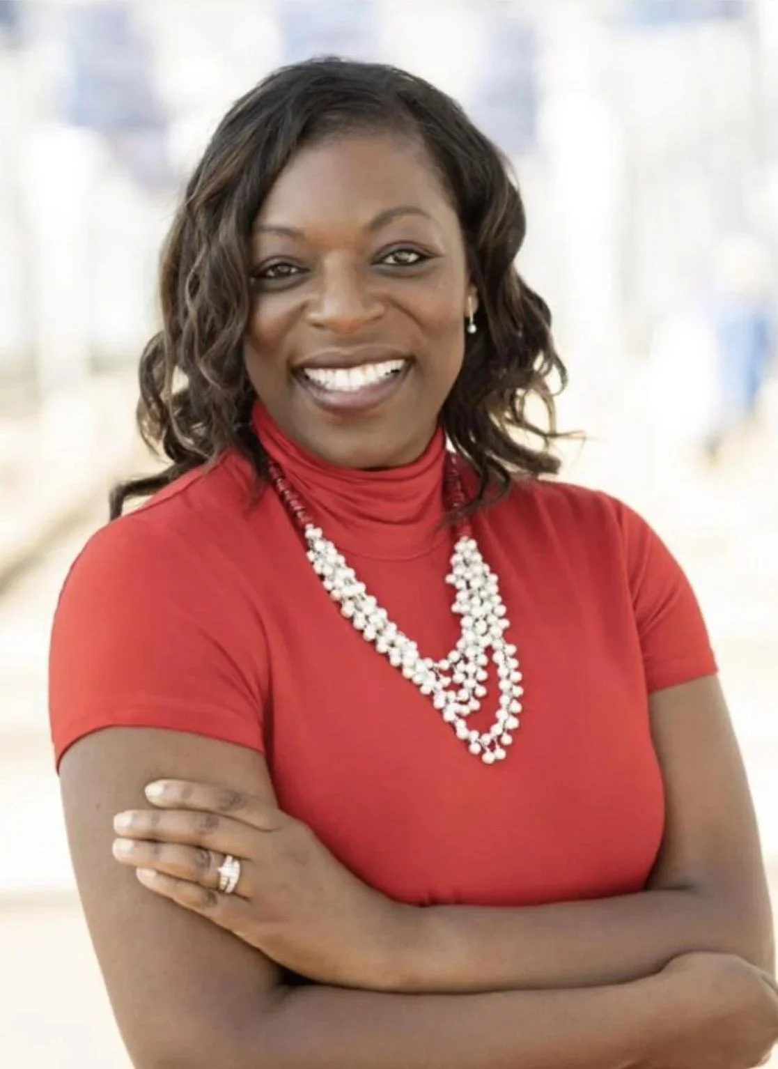 A woman with medium skin tone, dark wavy hair, wearing a red top, pearl necklace, and earrings, smiling with arms crossed.