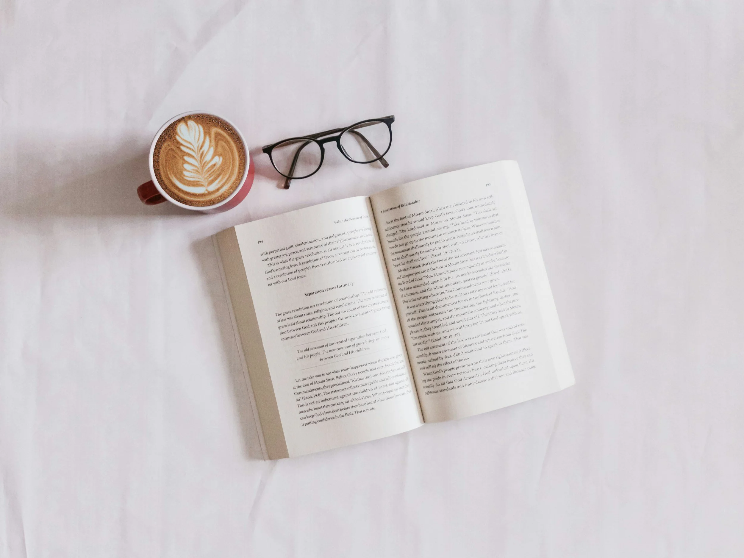 Open book, pair of glasses, and a cup of coffee with latte art on a white surface.