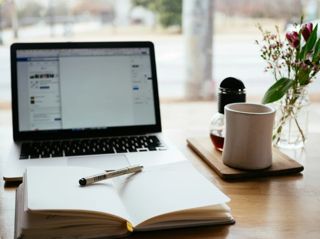 A workspace with an open notebook and pen, a laptop displaying a social media page, a white coffee mug, a plant with pink flowers, and a wooden tray holding a jar and a thermos, with a window in the background letting in natural light.