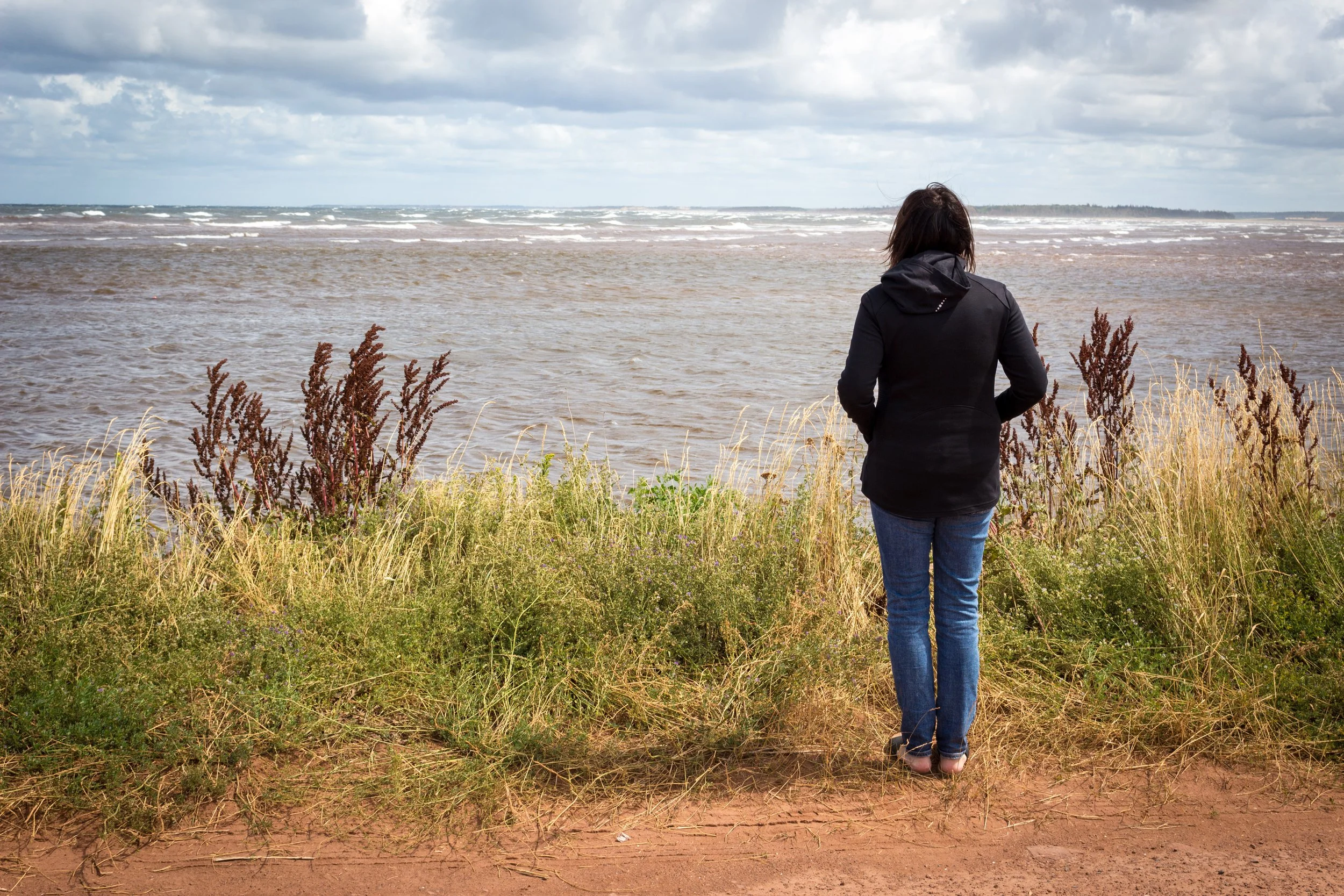 Woman in a black jacket and jeans looking at the ocean on a cloudy day, standing on a sandy beach with tall grass and plants in the foreground.