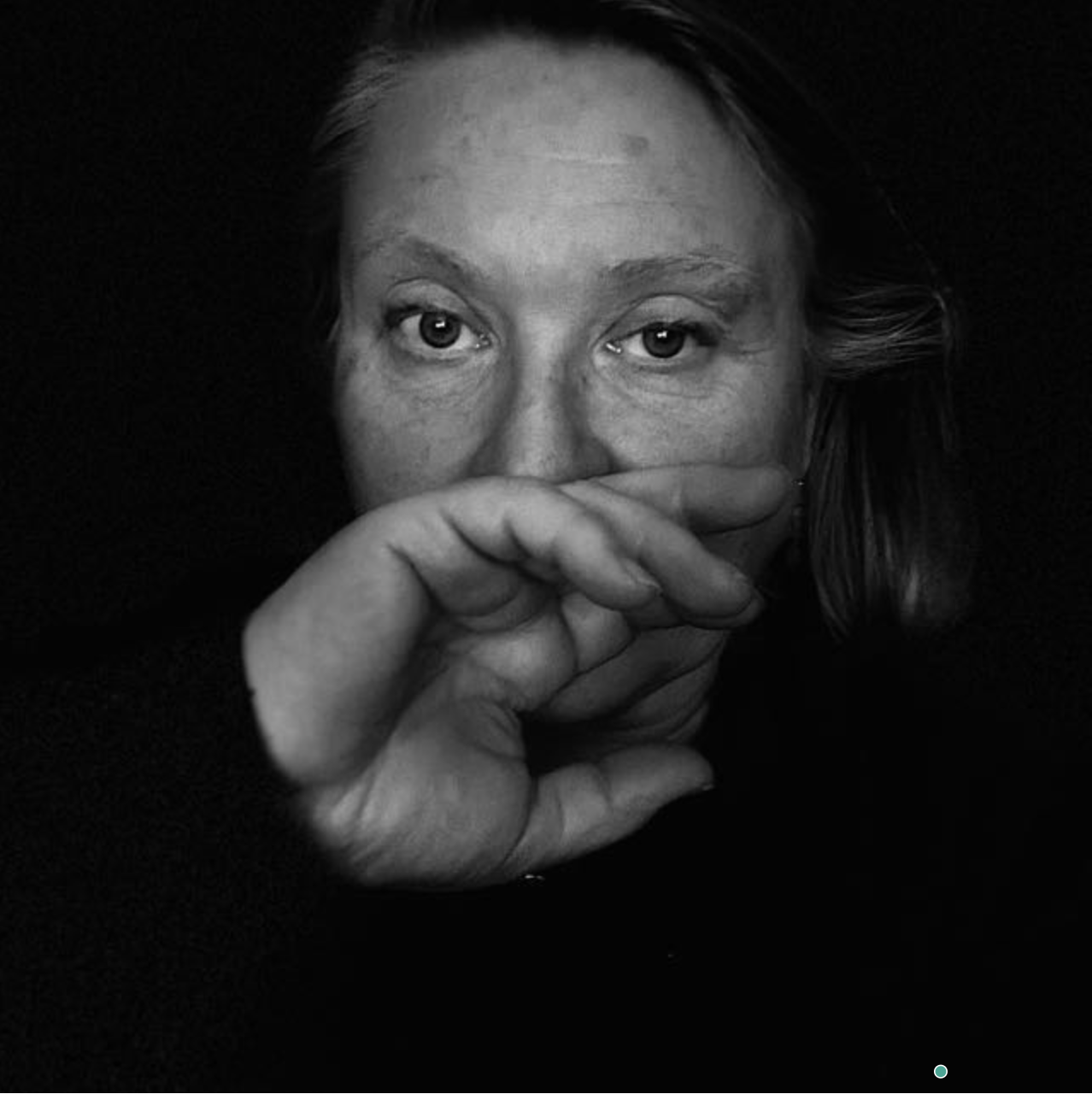 A black and white close-up portrait of a woman with her hand partially covering her face, looking directly into the camera.