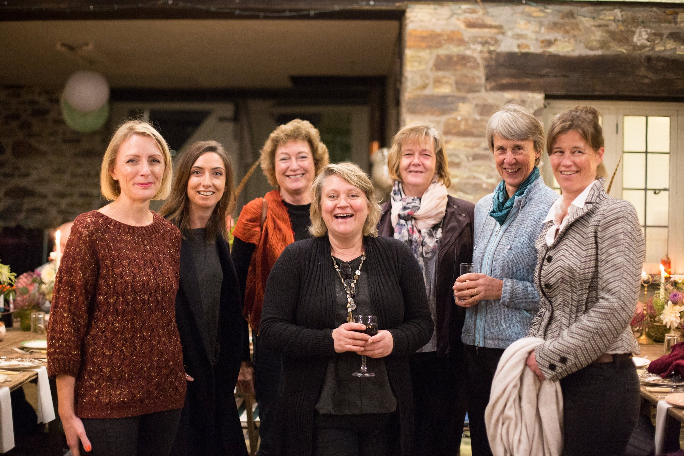 Group of seven women at a dinner party, smiling and holding drinks, in a cozy room with stone walls and warm lighting.