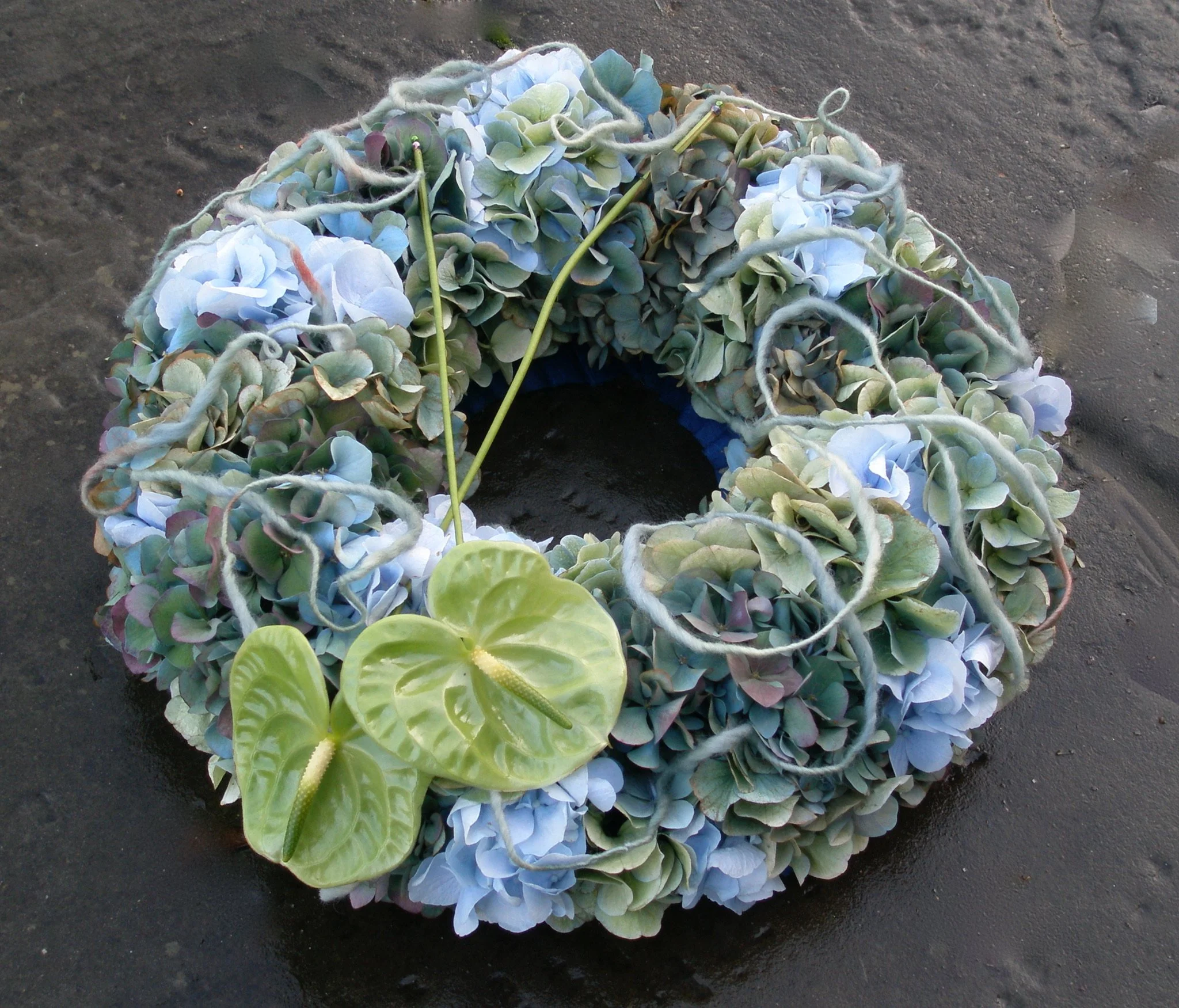 A floral arrangement made of pale blue and green hydrangea flowers, with an anthurium flower in the foreground, lying on dark sand or dirt.