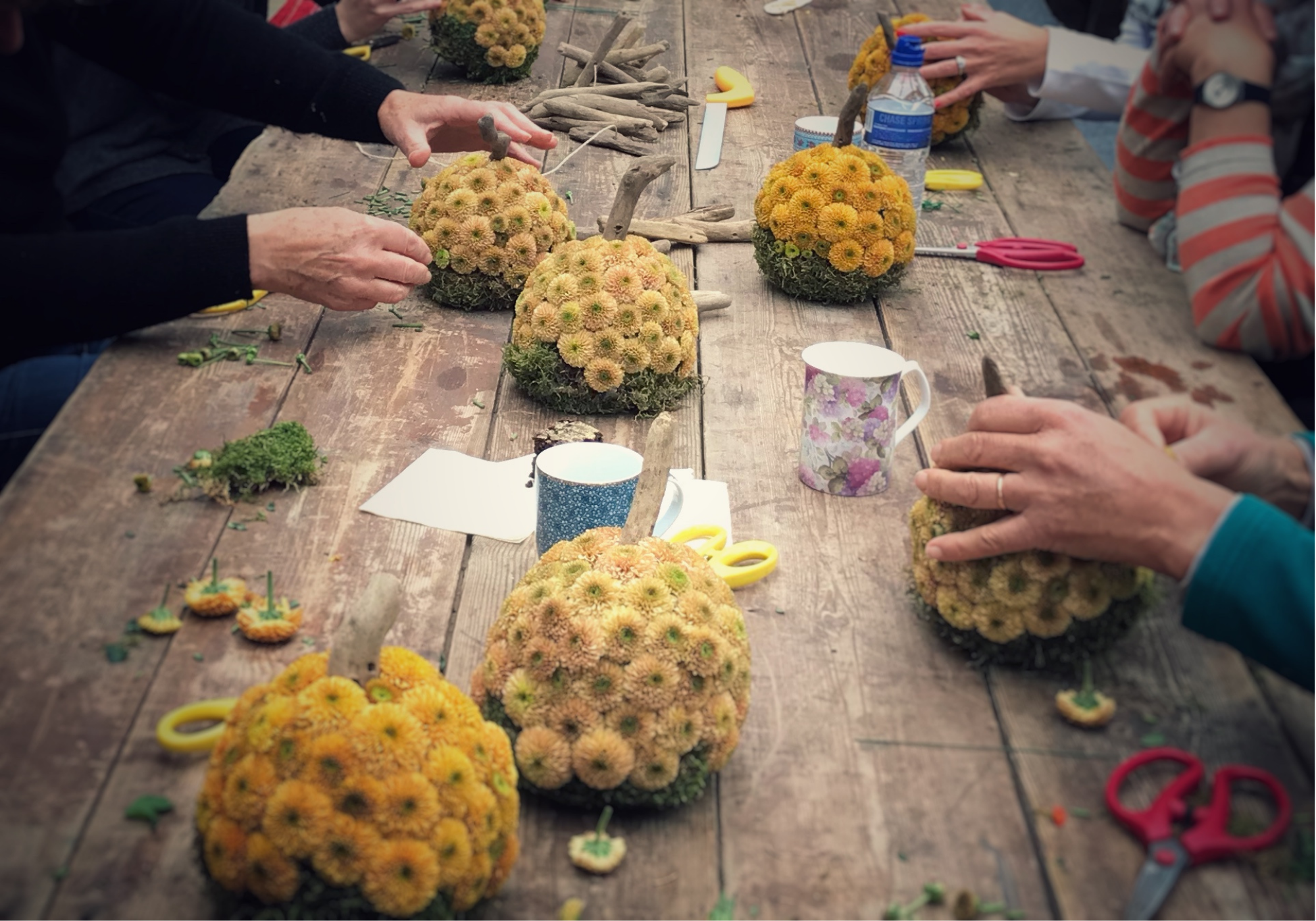 Several people working together to create floral arrangements using orange and yellow flowers on a wooden table, with scissors, glue, and other crafting tools scattered around.