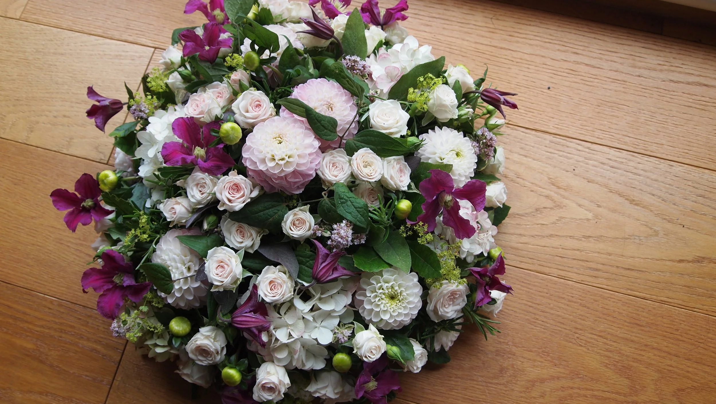 A floral bouquet with pink roses, white dahlias, purple and white hydrangeas, and greenery on a wooden floor.