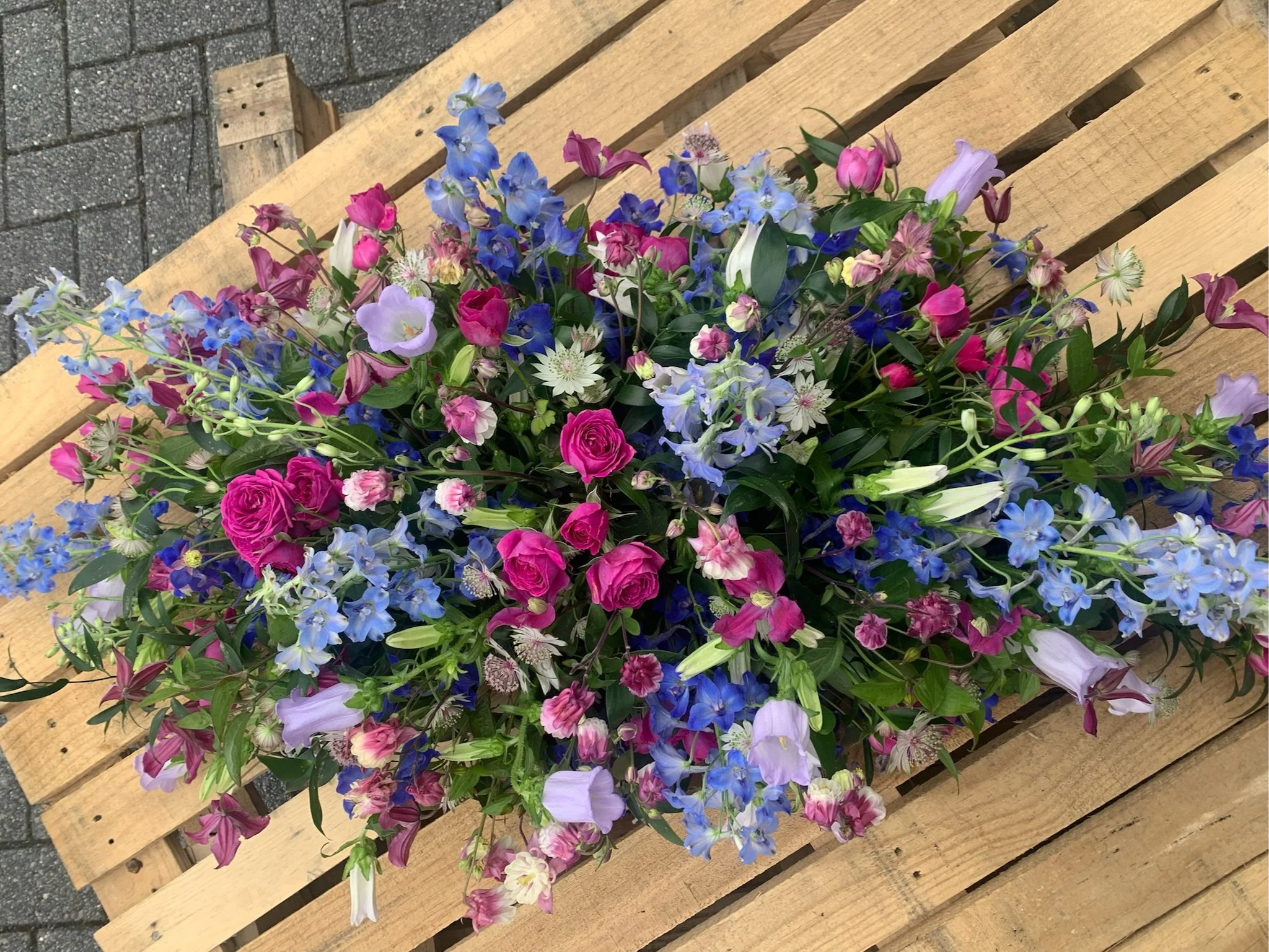 A wooden pallet with a colorful arrangement of flowers, including pink, purple, and blue blossoms, placed on a paved surface.