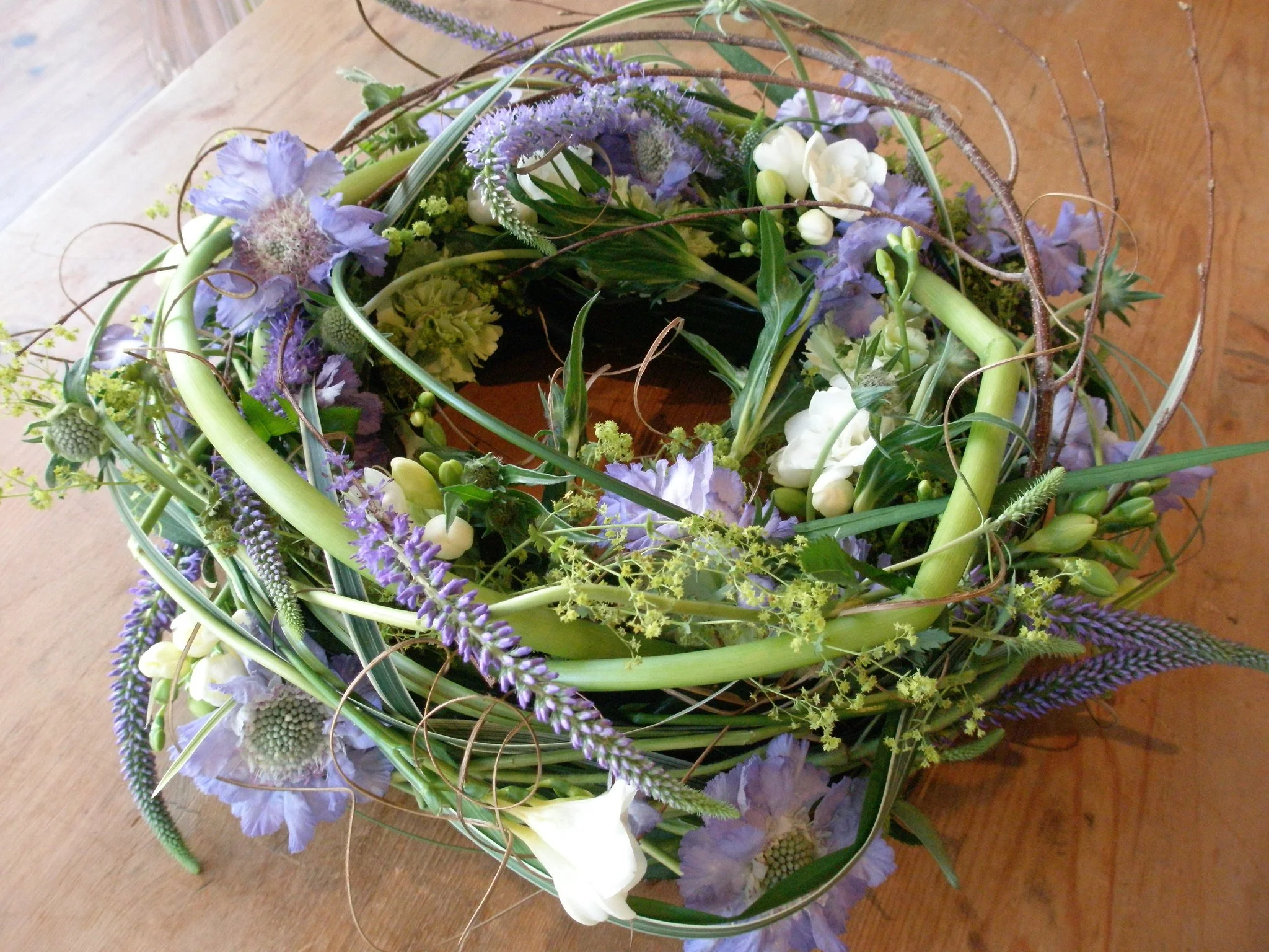 A floral arrangement with purple, white, and green flowers and greenery, placed on a wooden surface.