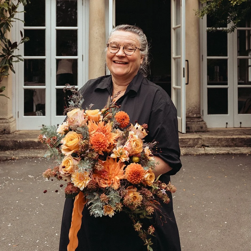 An elderly woman with glasses and gray hair smiling and holding a bouquet of orange and peach flowers in front of a building with white doors.