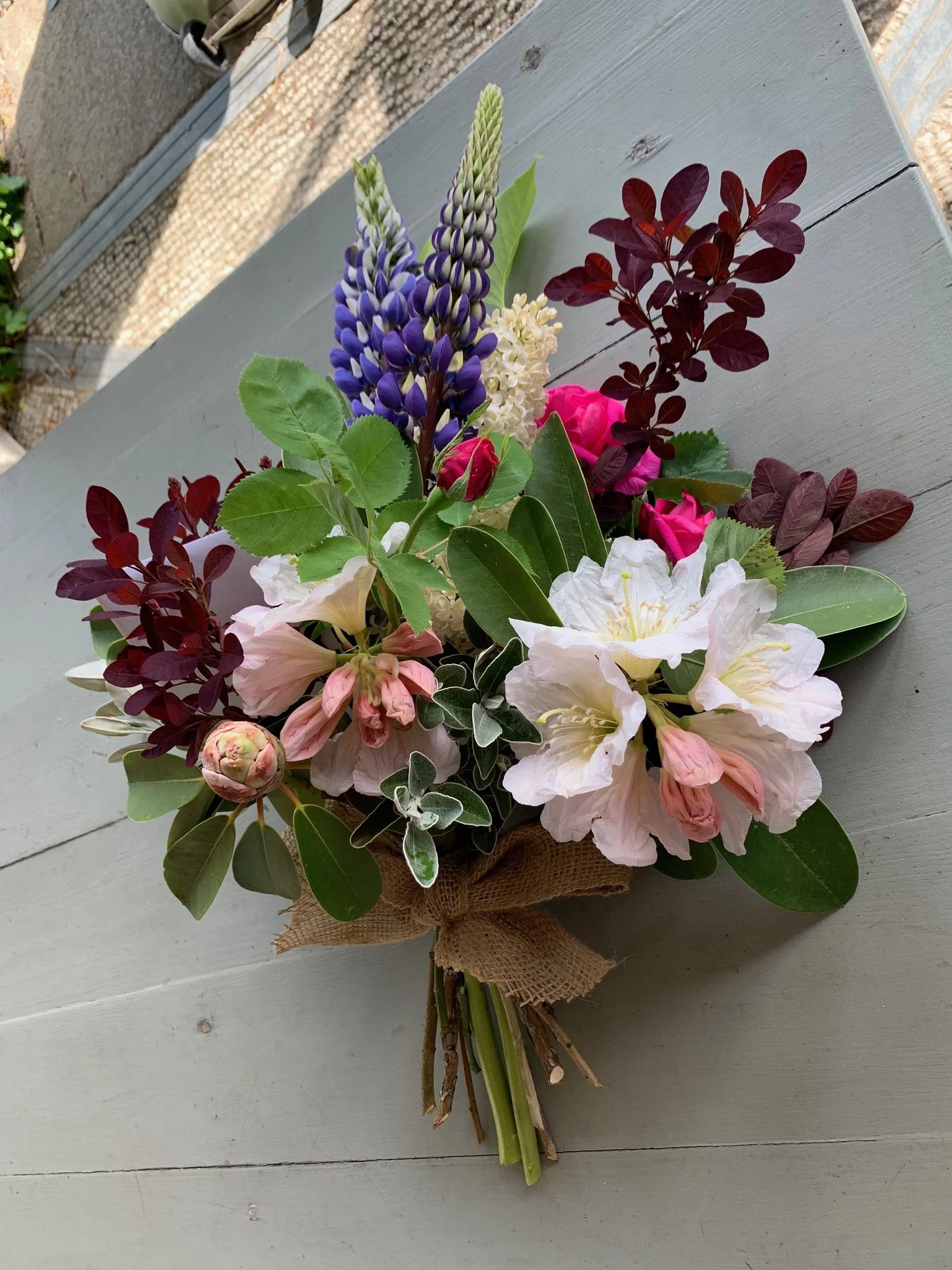 A colorful bouquet of flowers, including purple lupines, white and pink roses, white and pink azaleas, and dark red foliage, arranged on a wrapped bouquet on a light-colored wooden surface.