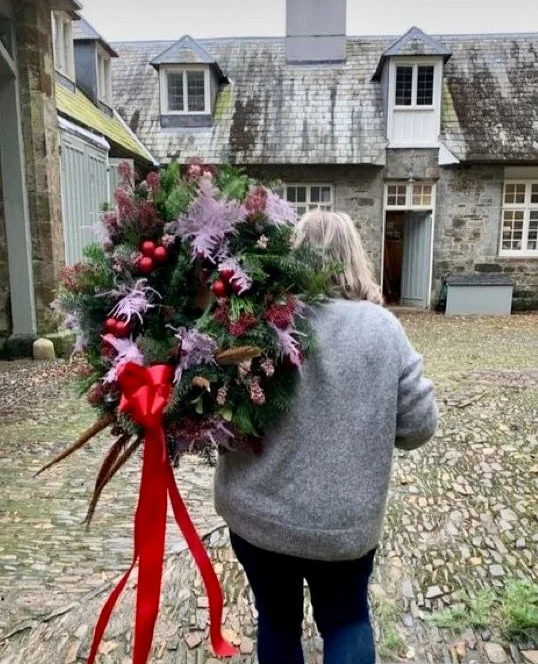 A person with gray hair and a gray coat walking away on a cobblestone path in front of a stone house, carrying a large Christmas wreath decorated with pine, pink and red ornaments, and a big red ribbon.
