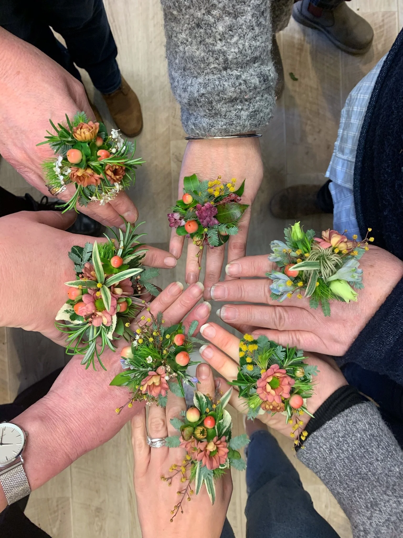 Multiple people wearing floral wrist corsages, with some touching or holding them, seen from above.