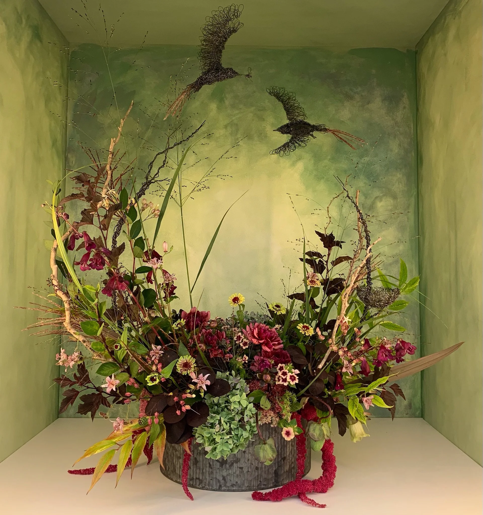 A large floral arrangement in a container with a striking display of various flowers, greenery, and decorative branches. The backdrop is a painterly green wall, emphasizing the vibrant and naturalistic composition.