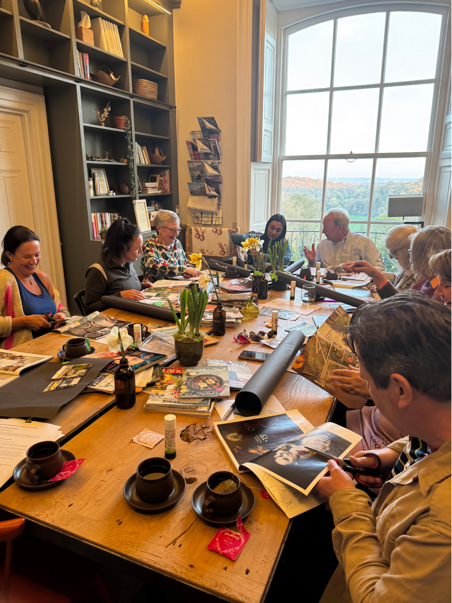 A group of people sitting around a large wooden table inside a room with a large bay window, several magazines and pamphlets, some plants, and bookshelves.