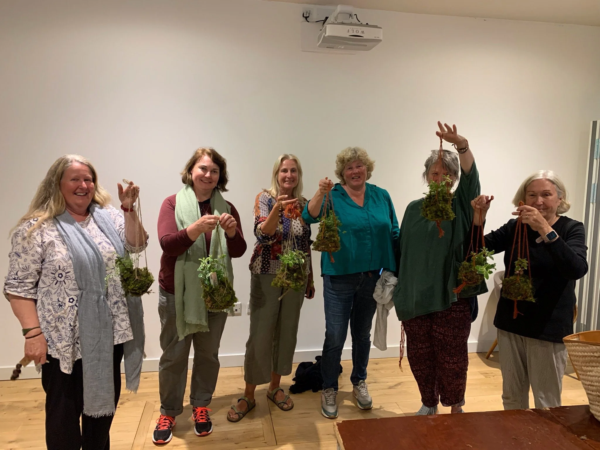 A group of seven women standing indoors, each holding hanging potted plants, smiling and posing for the photo.