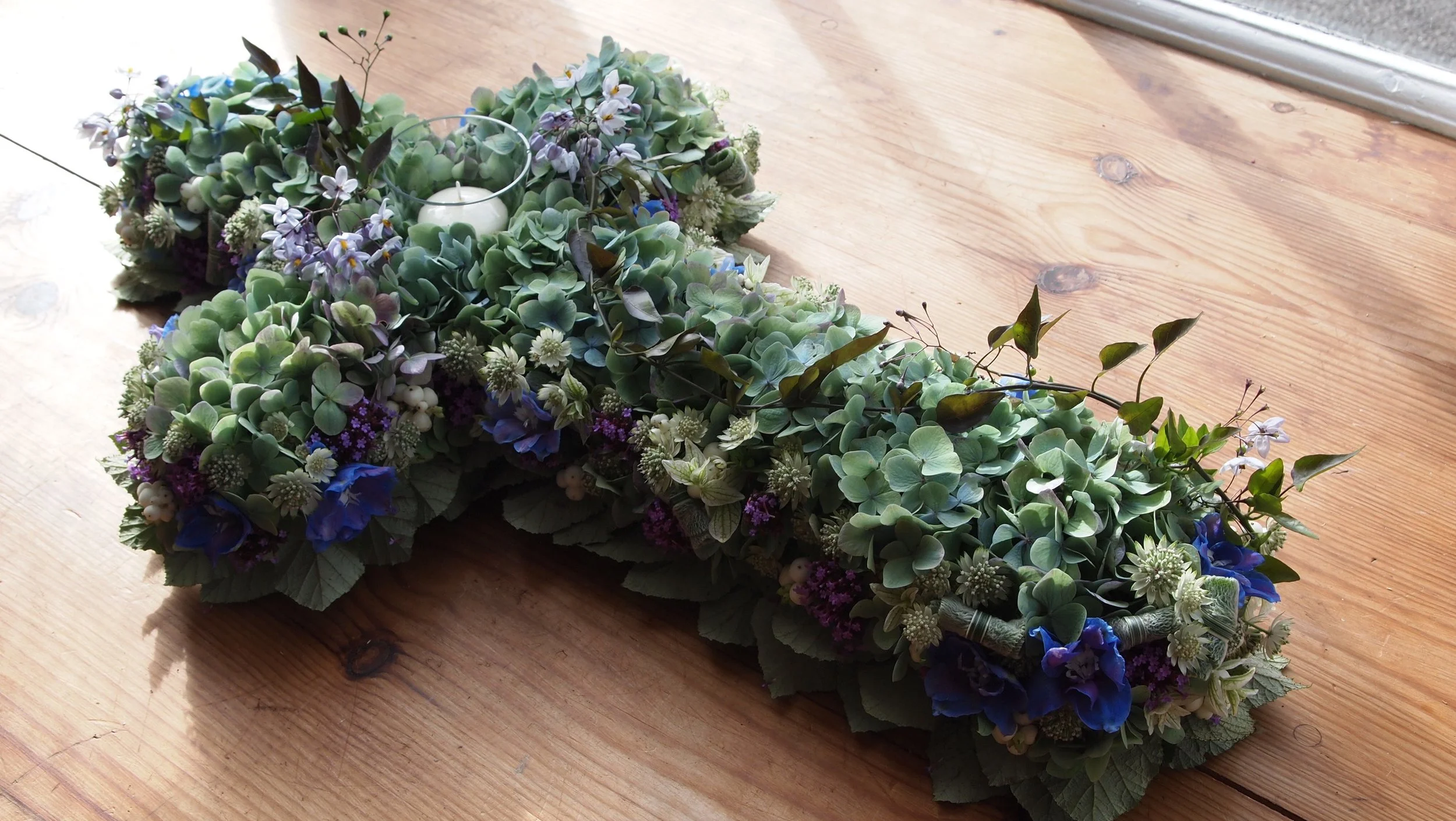 A floral arrangement with various green, white, purple, and blue flowers and leaves, featuring a small white candle, placed on a wooden surface near a window.