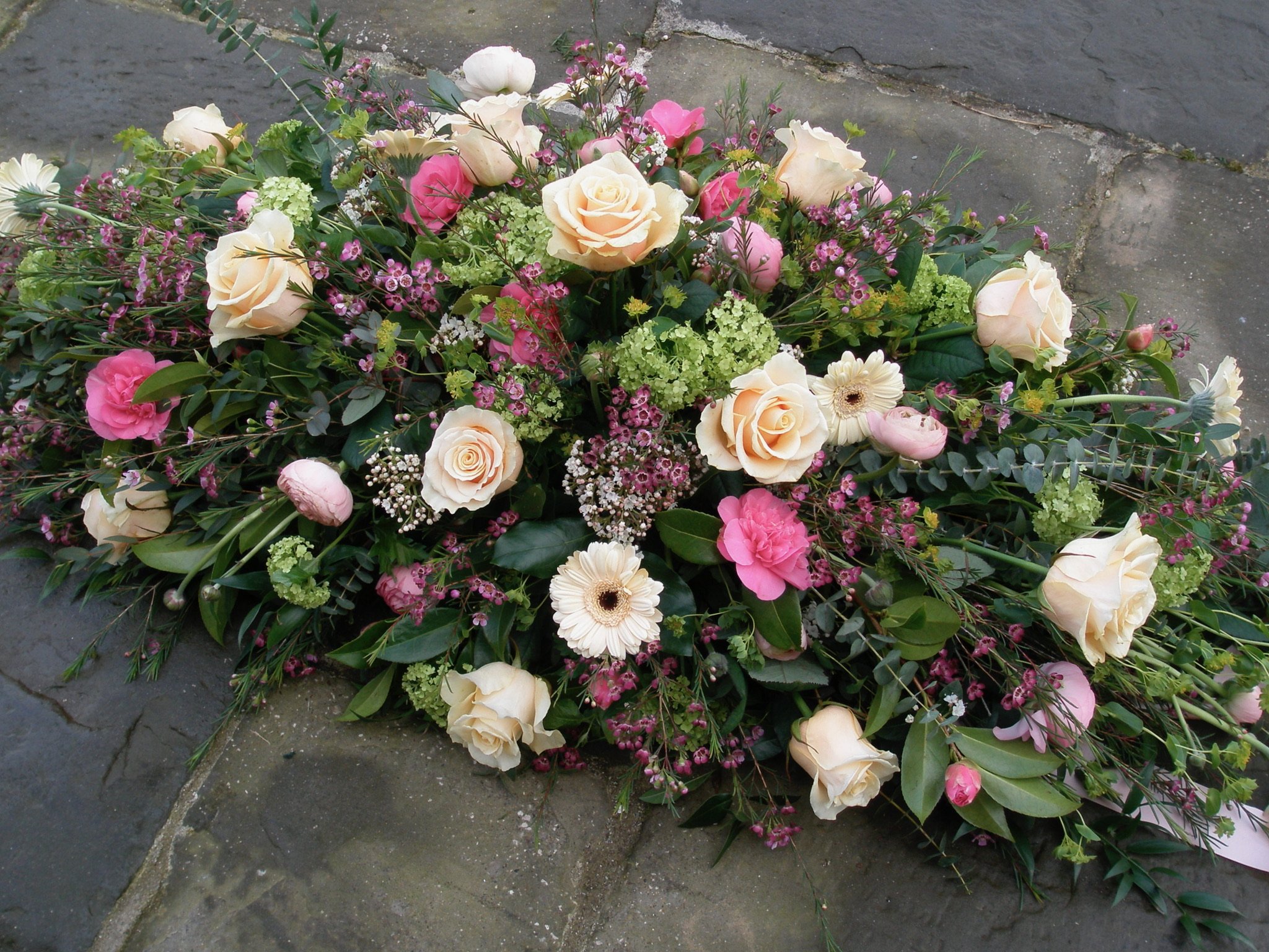 A floral arrangement on a pavement, featuring light-colored roses, pink flowers, white daisies, green hydrangeas, and various green foliage.
