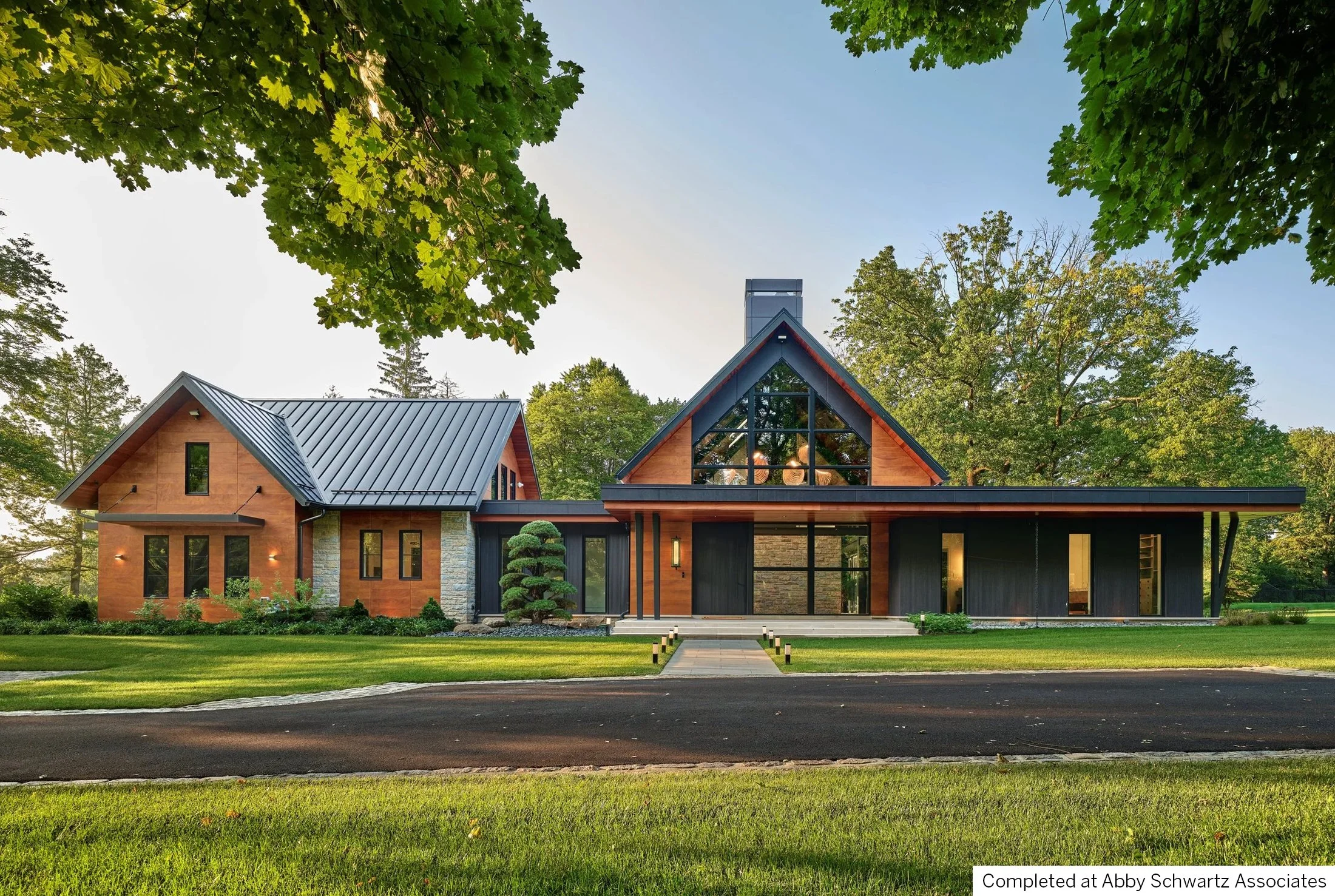 Modern, transitional, craftsman home with wooden and stone exterior, large glass windows, and a landscaped front yard with trees and a paved walkway.