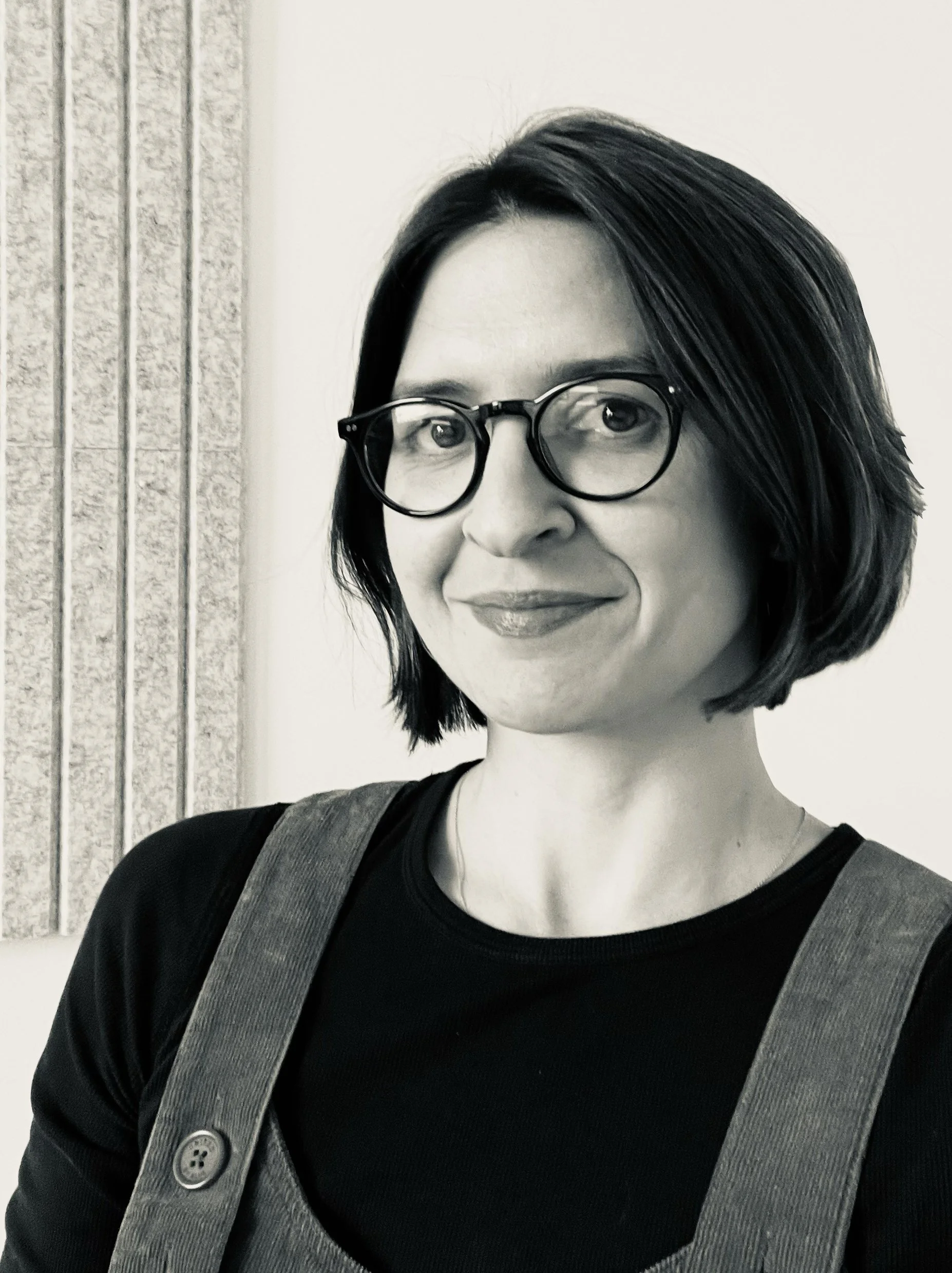 Black and white photo of a woman with short black hair, glasses, wearing a black top and overalls, smiling, standing indoors in front of a textured wall.