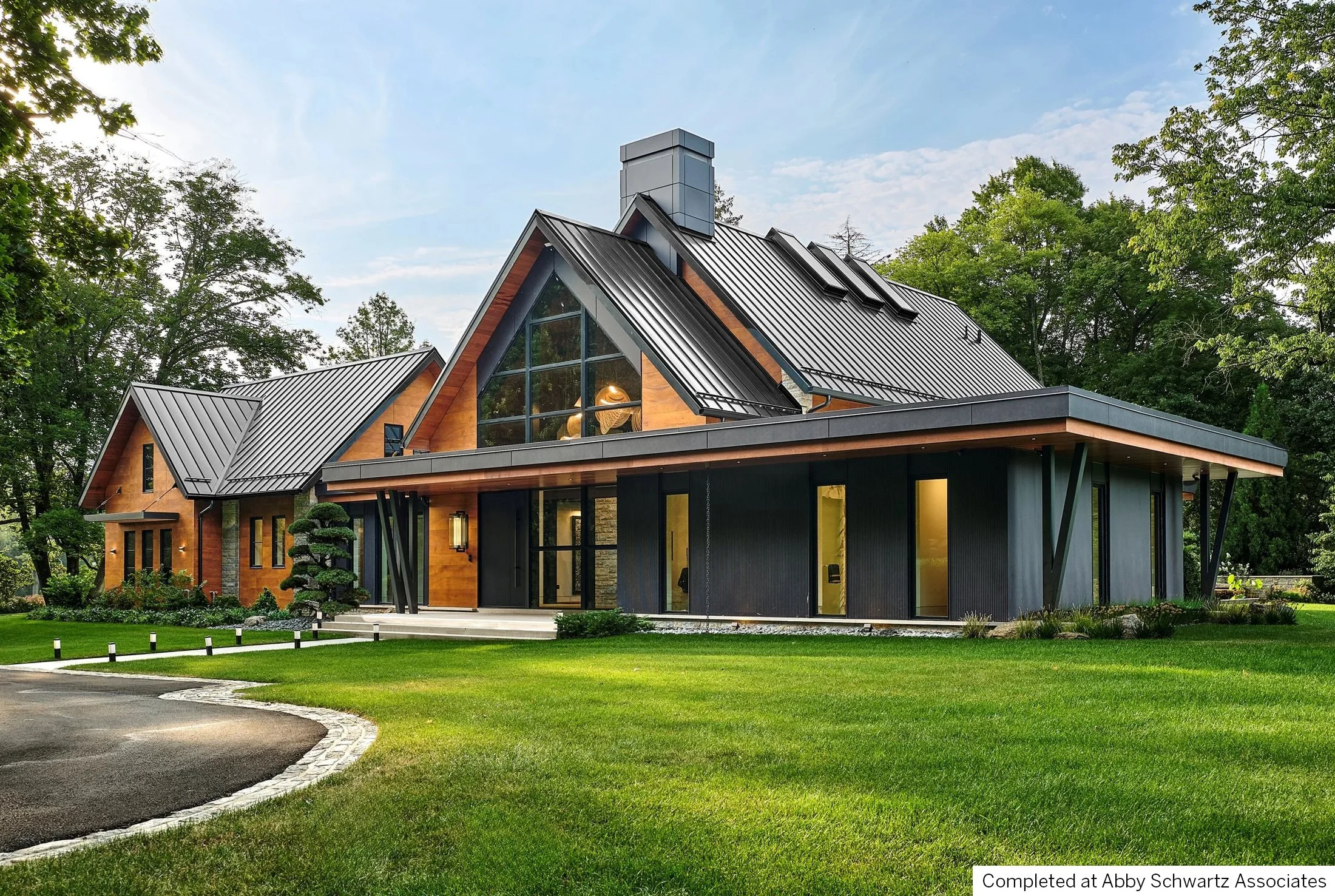 Warm modern, transitional, craftsman house with a combination of wooden and black exterior, large glass windows, and metal roofing, surrounded by a well-maintained lawn and trees.
