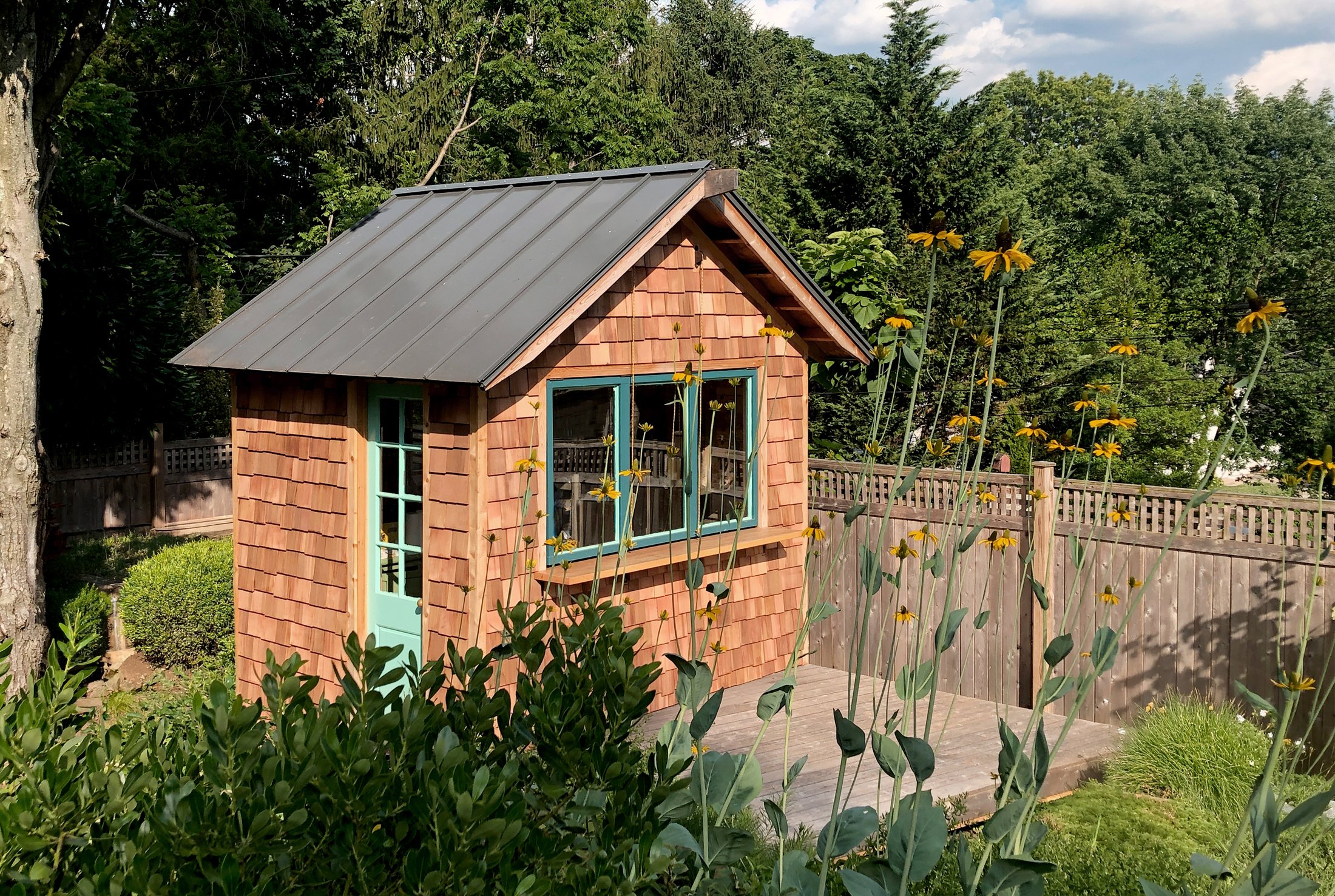 A small backyard shed with wooden shingle siding, a metal roof, and light blue window and door frames, surrounded by wildflowers and greenery, with a wooden fence in the background.