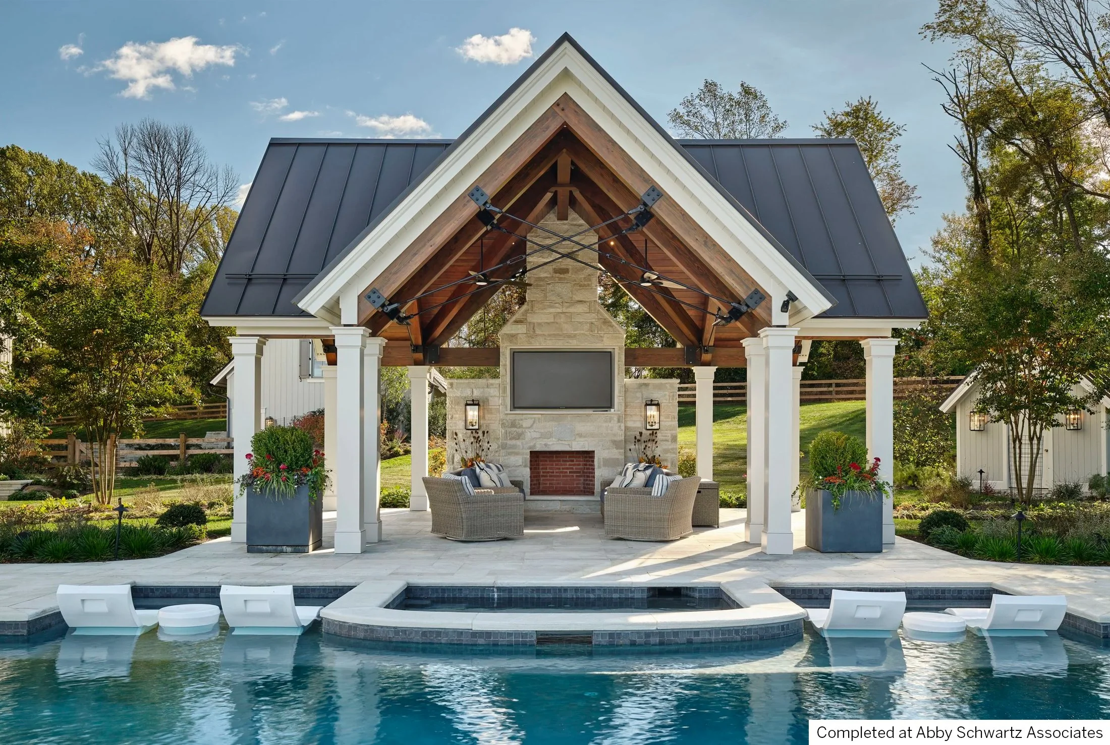 A backyard with a pool in front of a covered outdoor living area featuring a stone fireplace, a large television, chairs, and potted plants, all under a gabled roof with black metal panels.