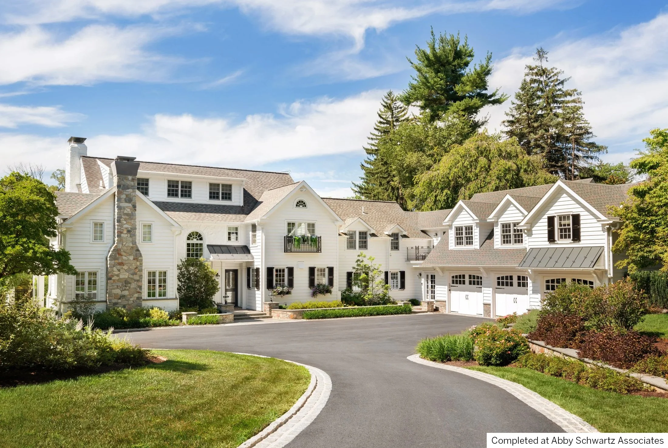 A large, traditional white house with multiple gabled roofs, a stone chimney, and a paved driveway surrounded by green grass and trees.
