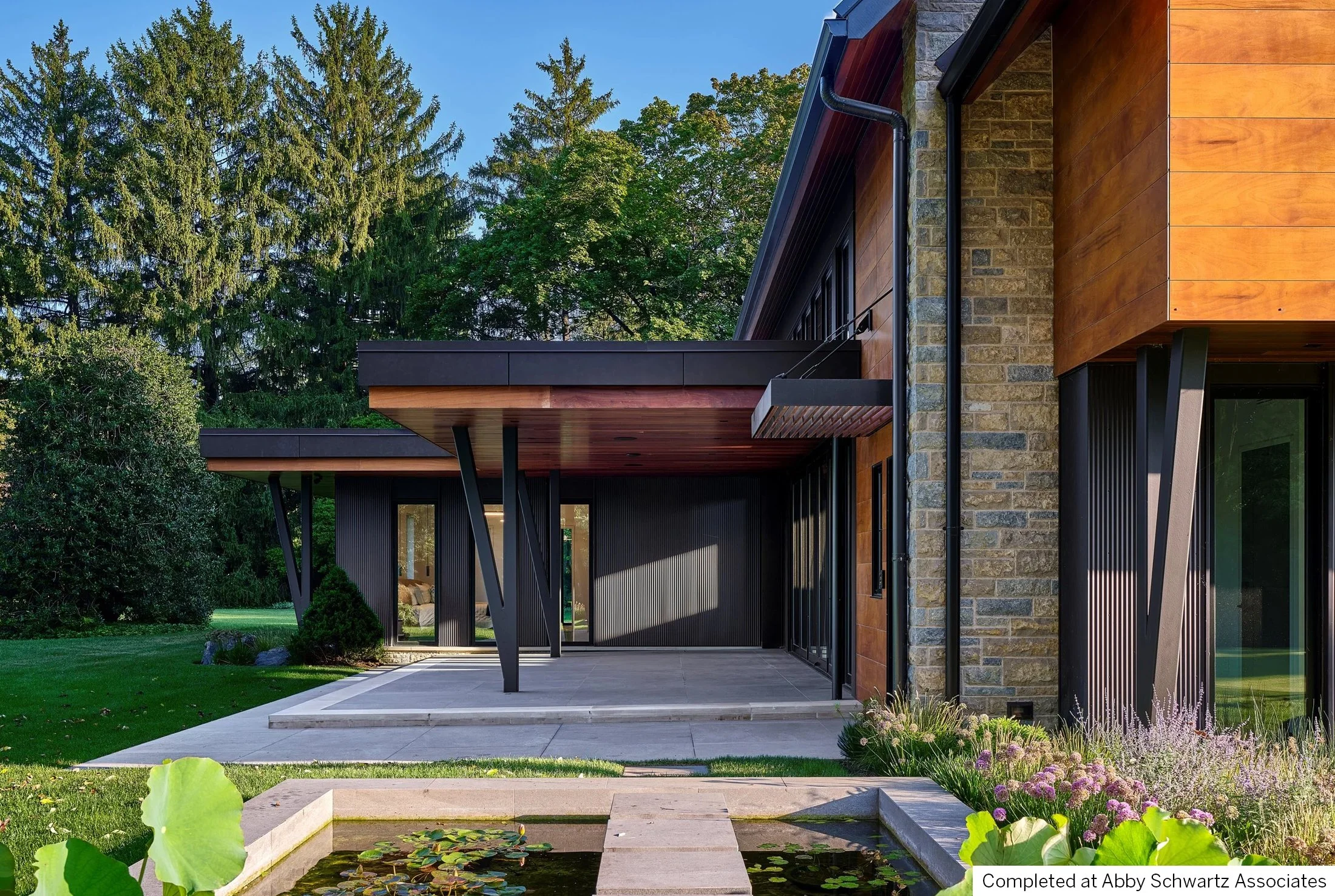 Modern house with black metal columns, wooden siding, and stone accents, surrounded by lush green trees and a small pond with lily pads in the foreground.