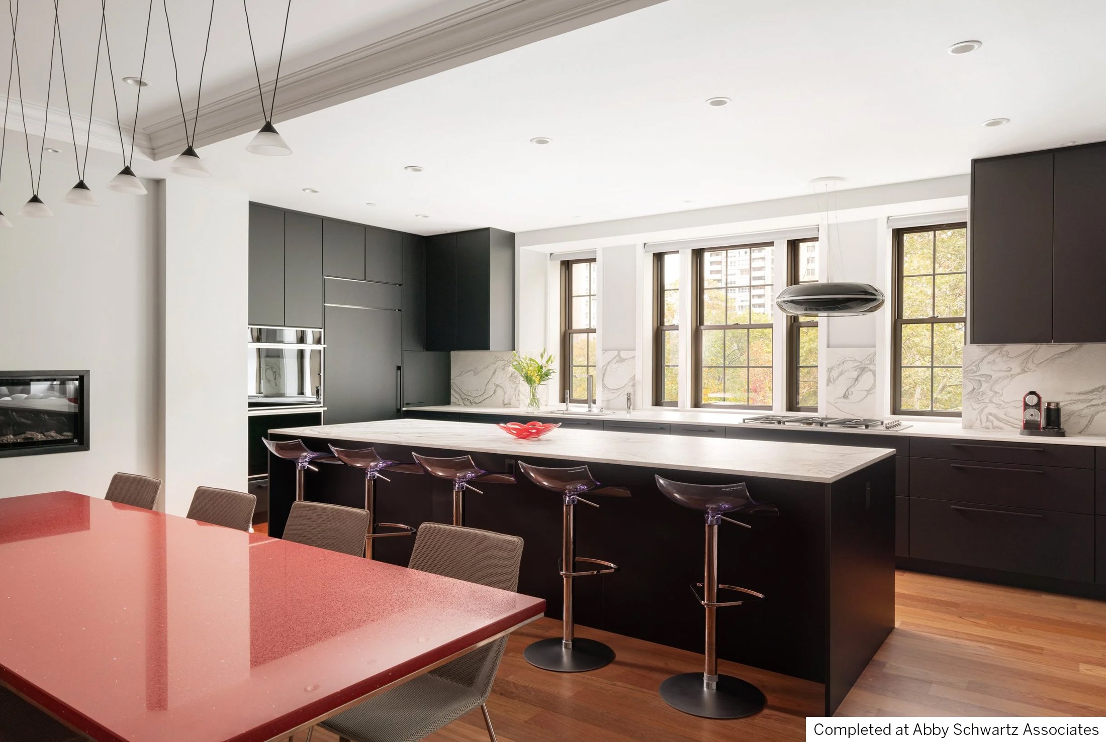 Modern kitchen with white marble island, black cabinets, large windows, pendant light, and red dining table with beige chairs.