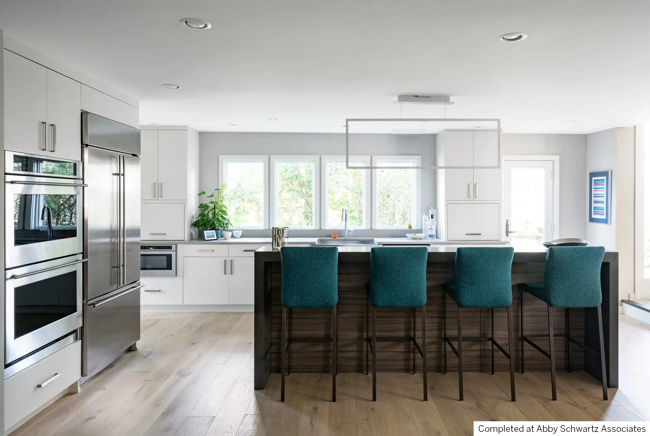 Modern kitchen with white cabinets, stainless steel appliances, a dark wood island with four teal chairs, large windows, and minimal decor.