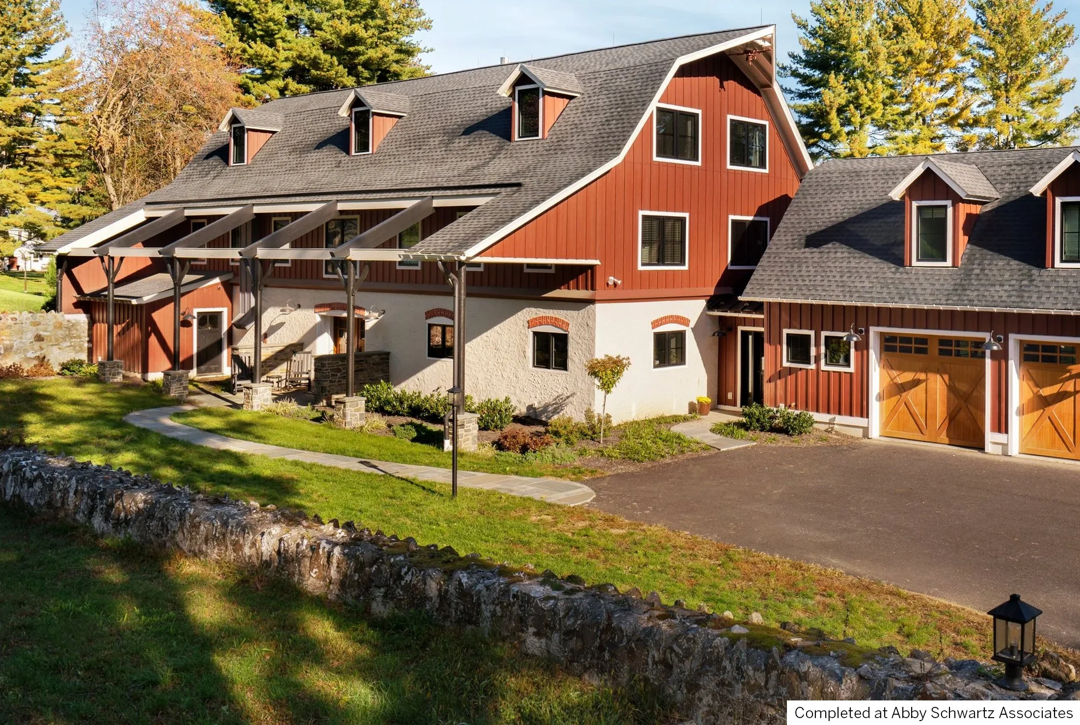 A large, modern farmhouse home with a red exterior and wooden accents, surrounded by green grass and trees, featuring a front courtyard with a walkway, small plants, and a driveway with a garage.