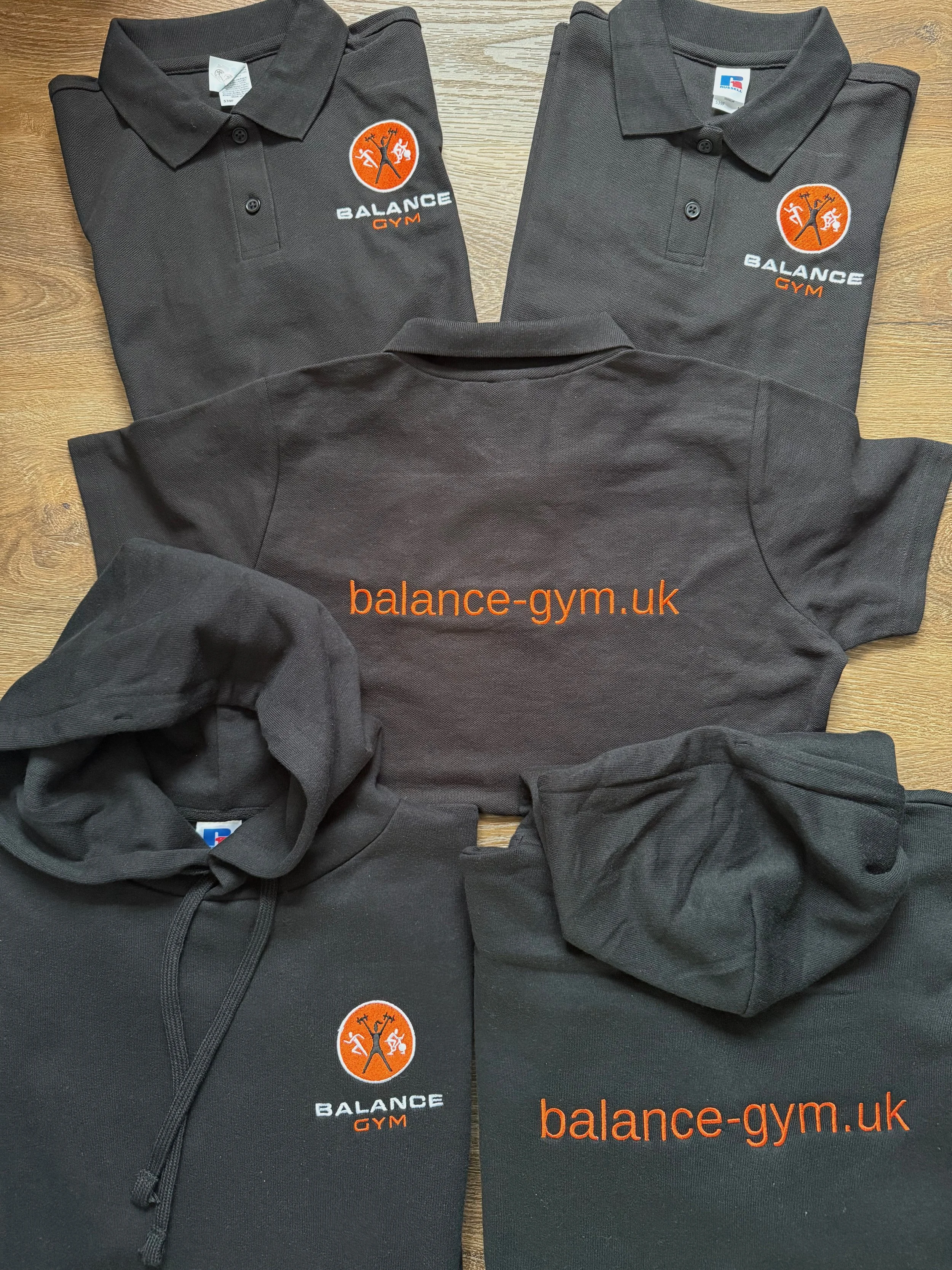 Two polo shirts, a hoodie, and a sweatshirt with logos and website text related to Balance Gym, placed on a wooden surface.