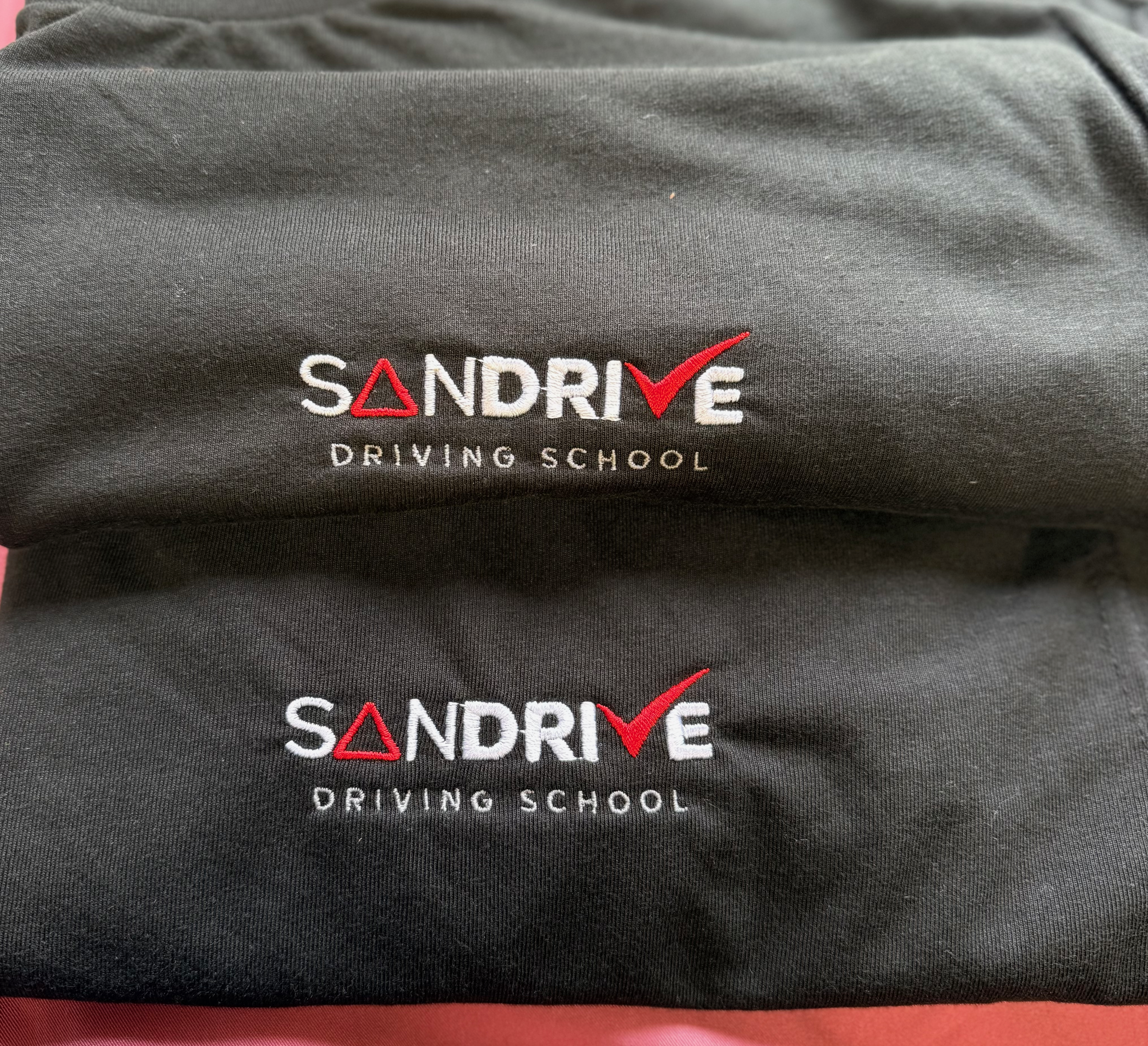 Two black cloth bags with embroidered 'SANDRIVE DRIVING SCHOOL' logos, featuring red checkmarks, one above the other.