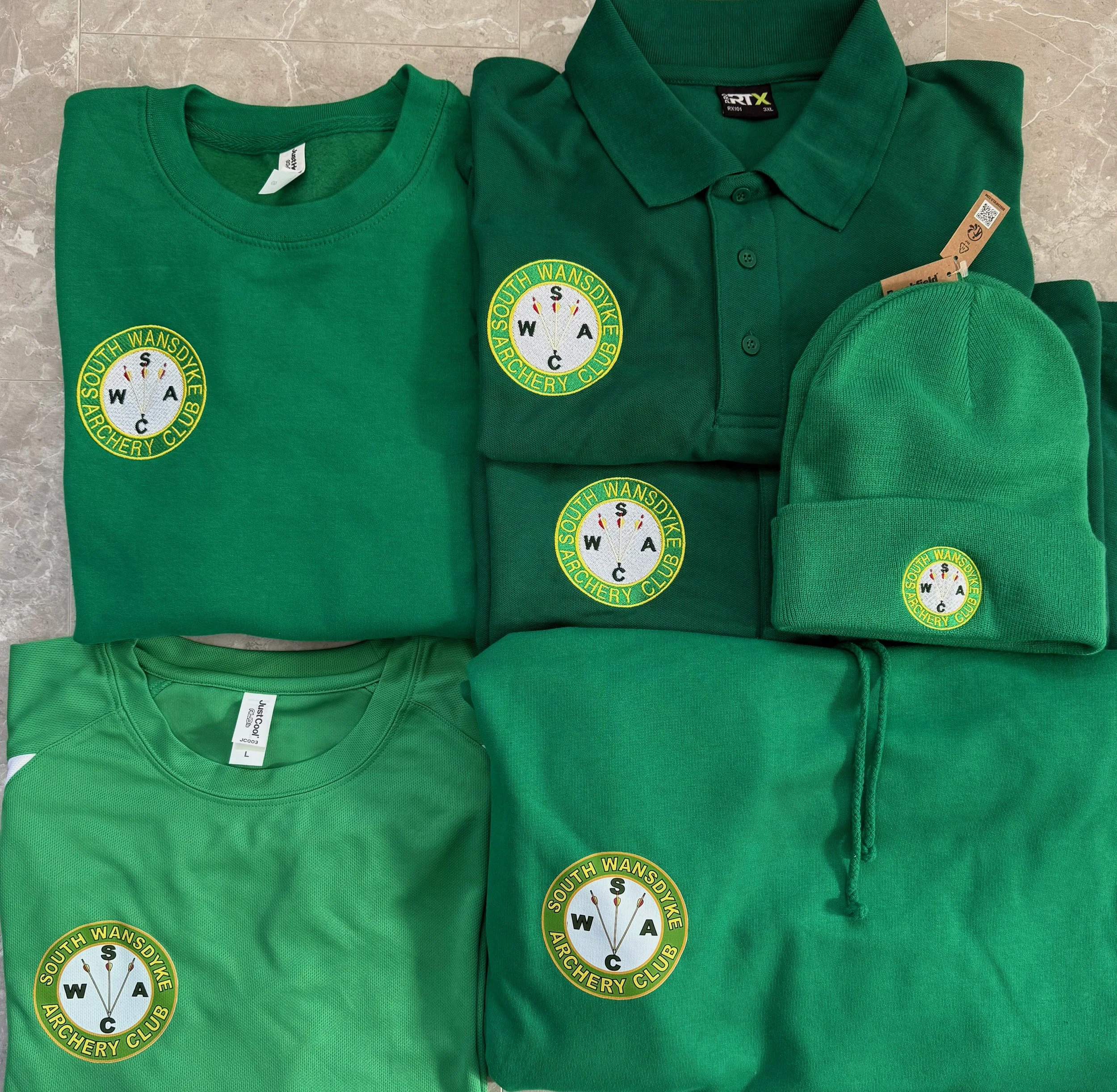 A collection of green clothing items, including shirts, a hoodie, a beanie, and a polo shirt, all with the embroidered logo of the South Wansdyke Archery Club. The logo features a circular design with a compass needle and the club name.